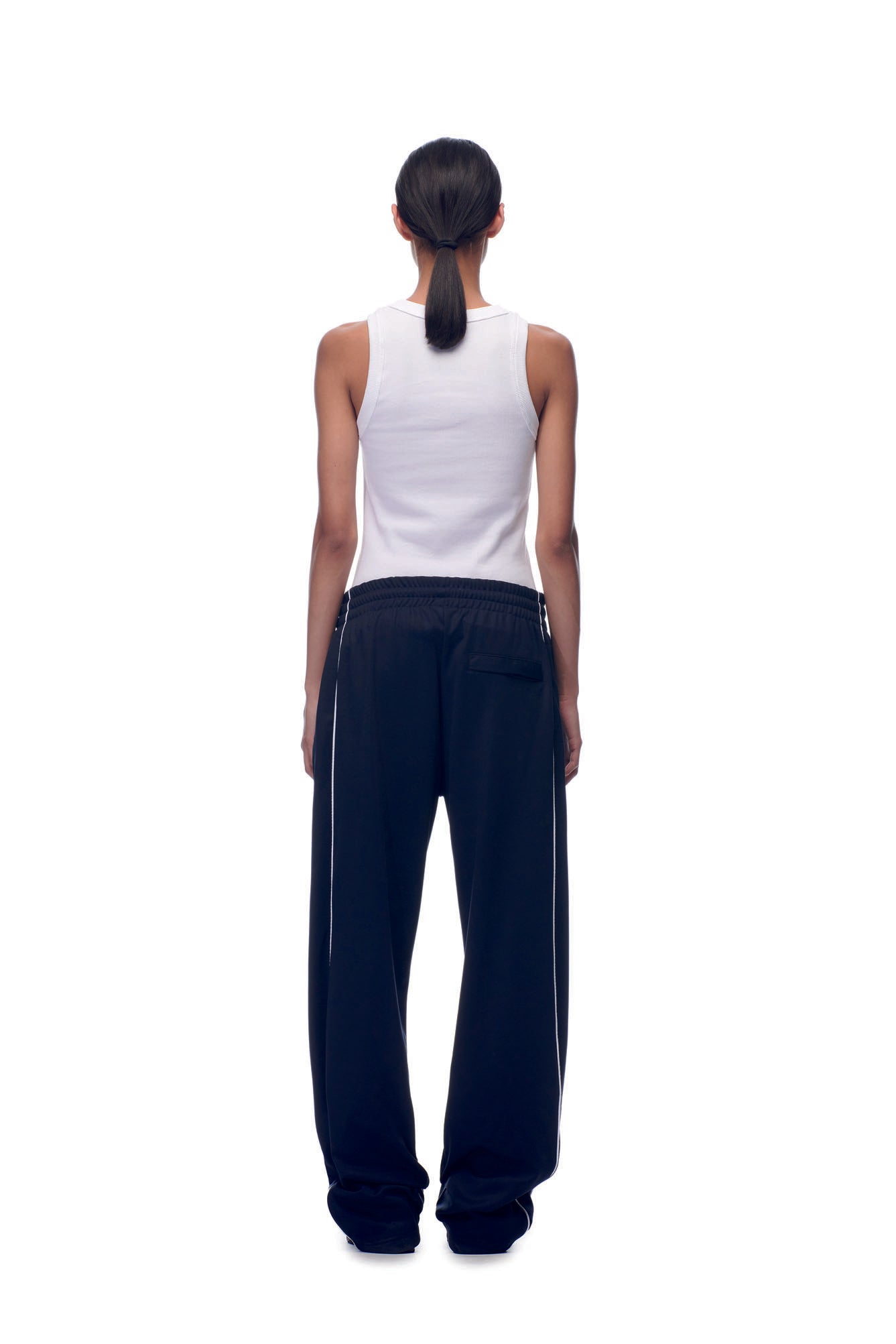 A person with long dark hair tied back, dressed in a white sleeveless top and 6PM TENNIS TRAINER PANTS BLUE, stands facing away against a plain white background.