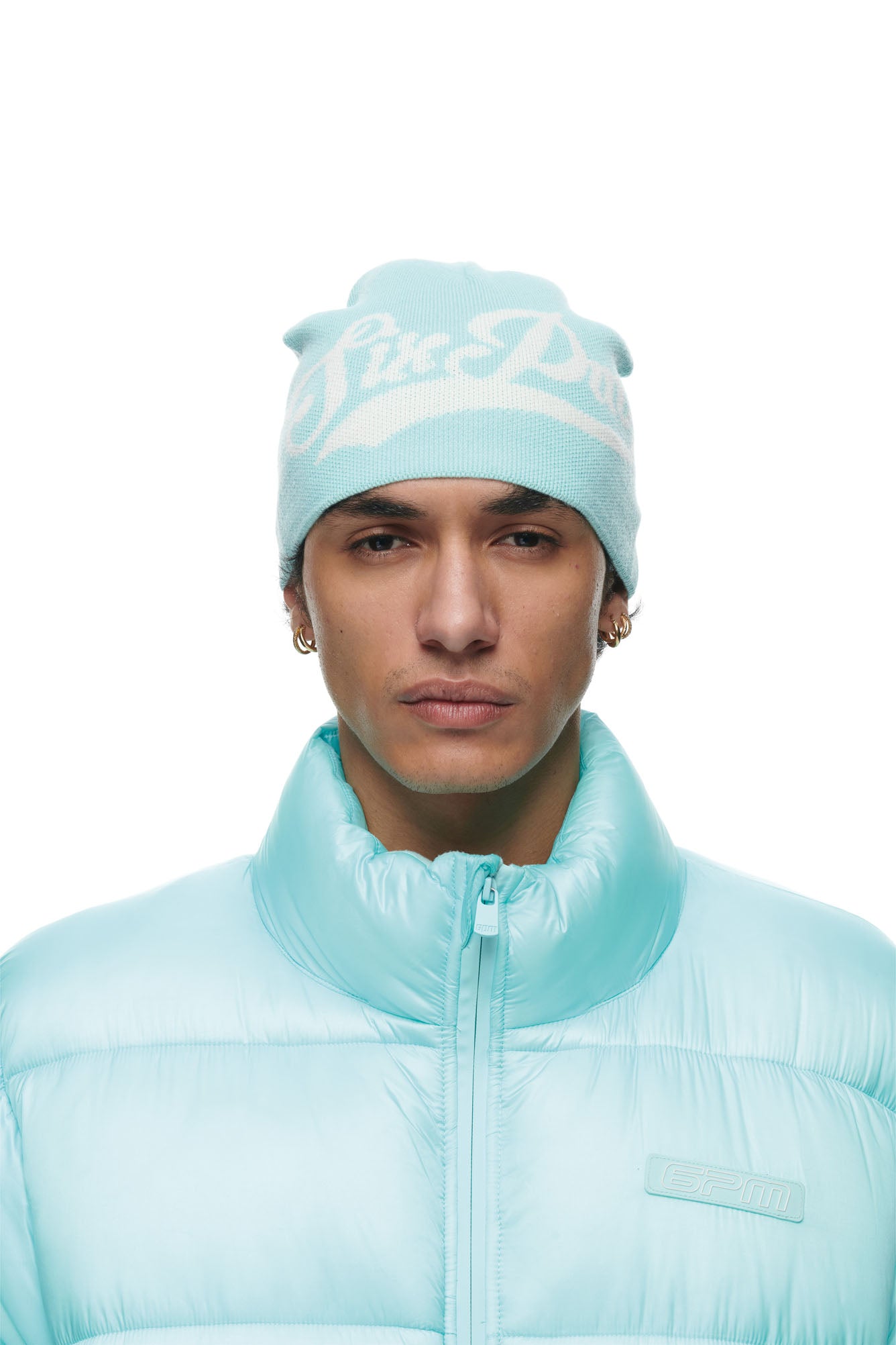 A person wearing a turquoise 6PM SCRIPT LOGO BEANIE with white lettering and a light blue puffer jacket stands against a plain white background.