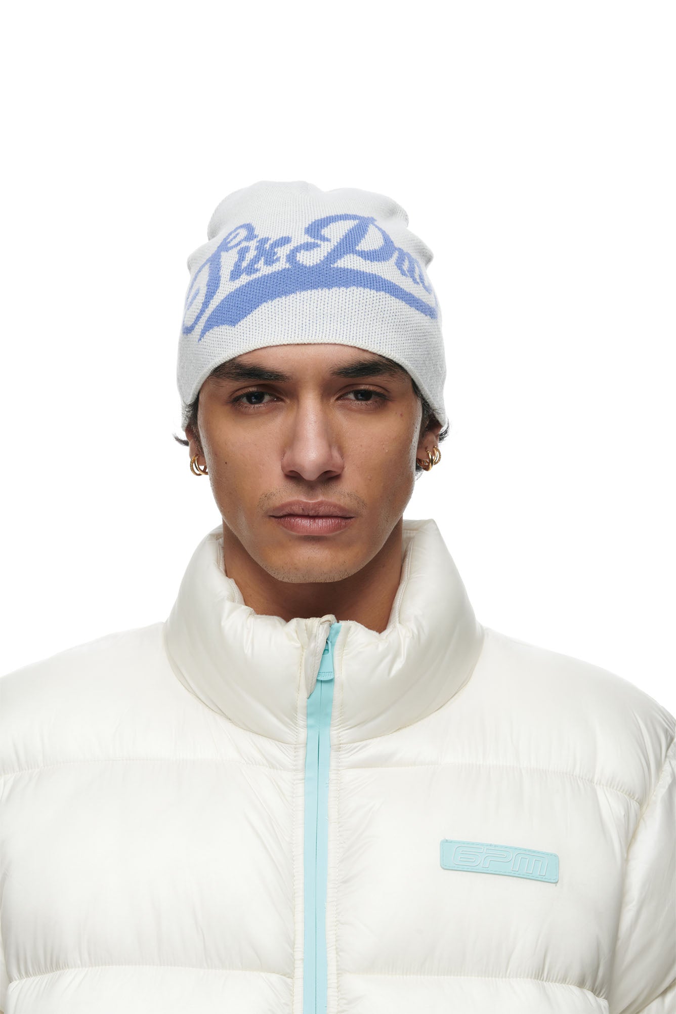 A person wears the 6PM SCRIPT LOGO BEANIE CREAM and a white puffer jacket with a light blue zipper and logo, looking directly at the camera with a neutral expression.