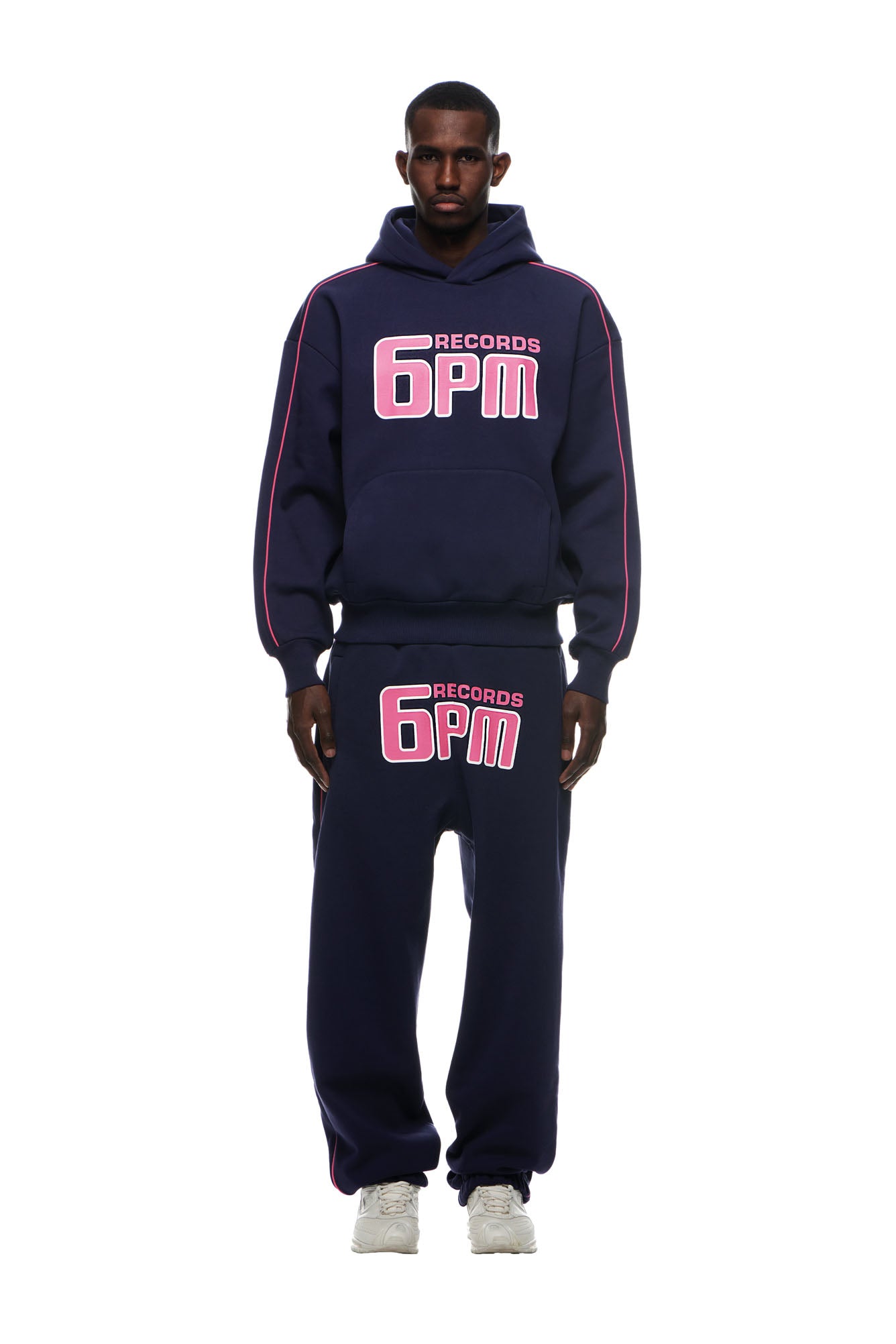 Someone stands against a white background wearing the 6PM RECORDS HOODIE DEEP BLUE and matching sweatpants, both featuring bold pink and white 6PM RECORDS lettering, paired with white sneakers.