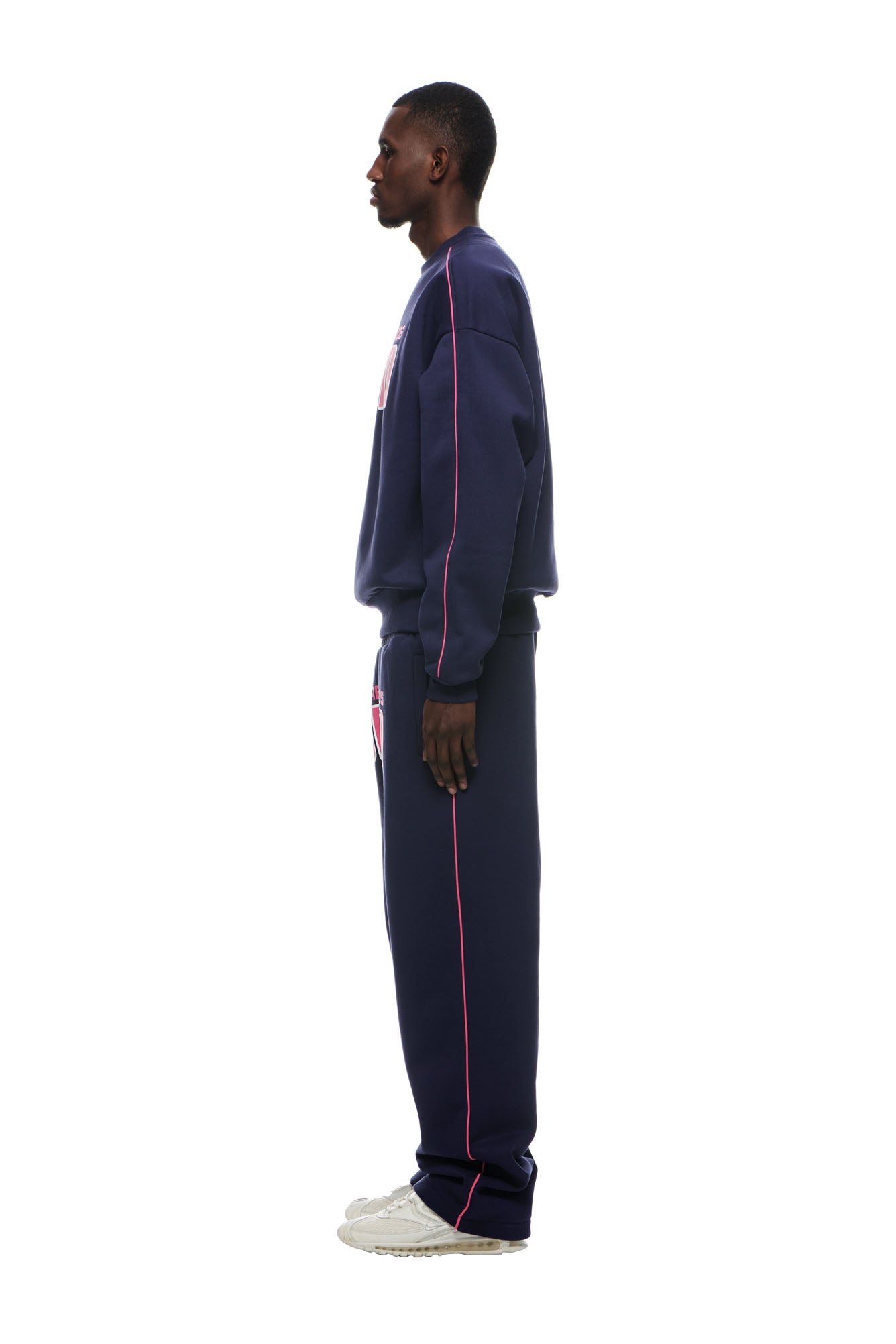 A man in profile faces left, wearing the 6PM RECORDS CREWNECK DEEP BLUE sweatshirt and matching pants with pink piping, plus white sneakers, against a plain white background.