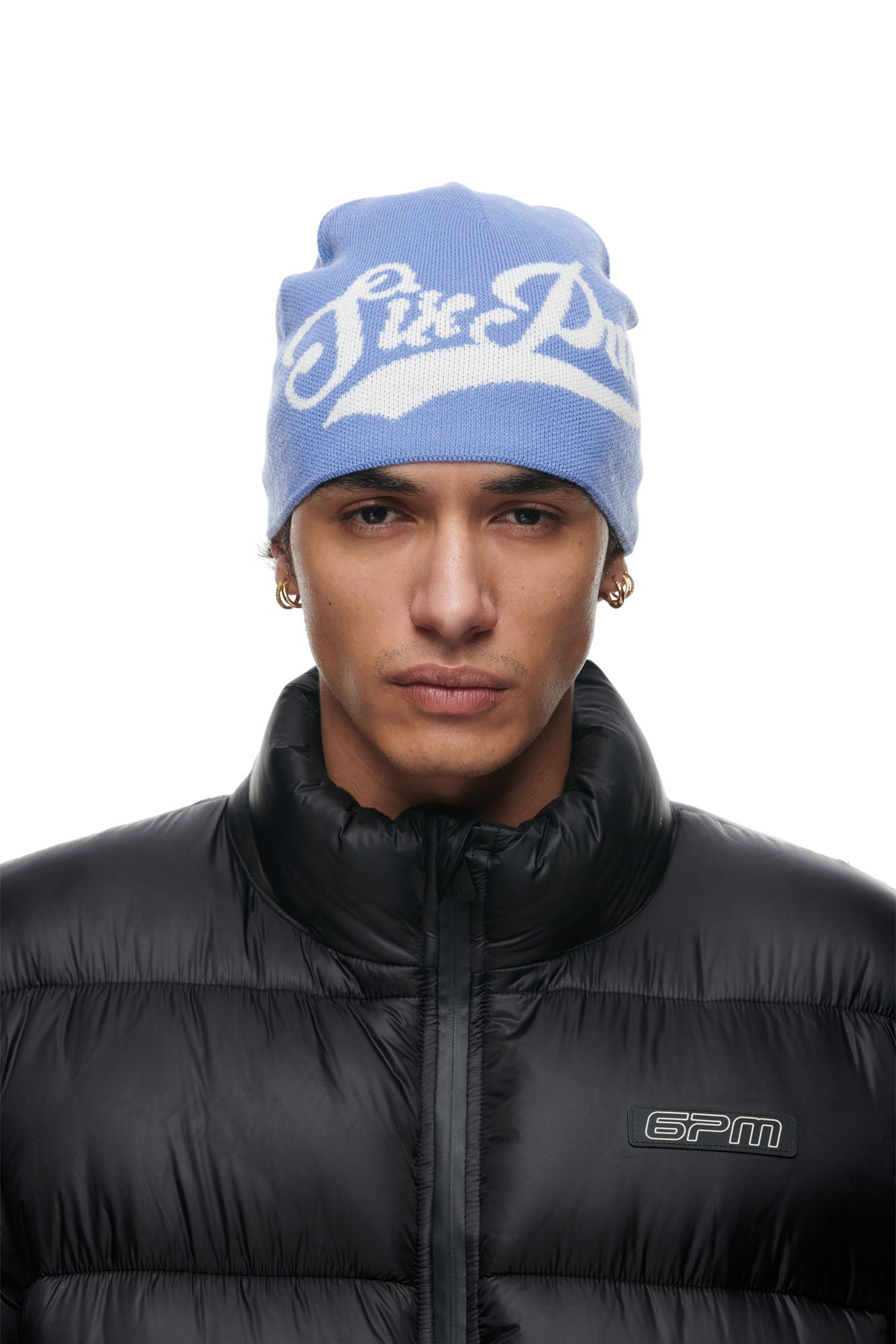 A person wearing the 6PM SCRIPT LOGO BEANIE in Persian Blue, a black puffer jacket, and gold hoop earrings stands facing forward against a plain white background.