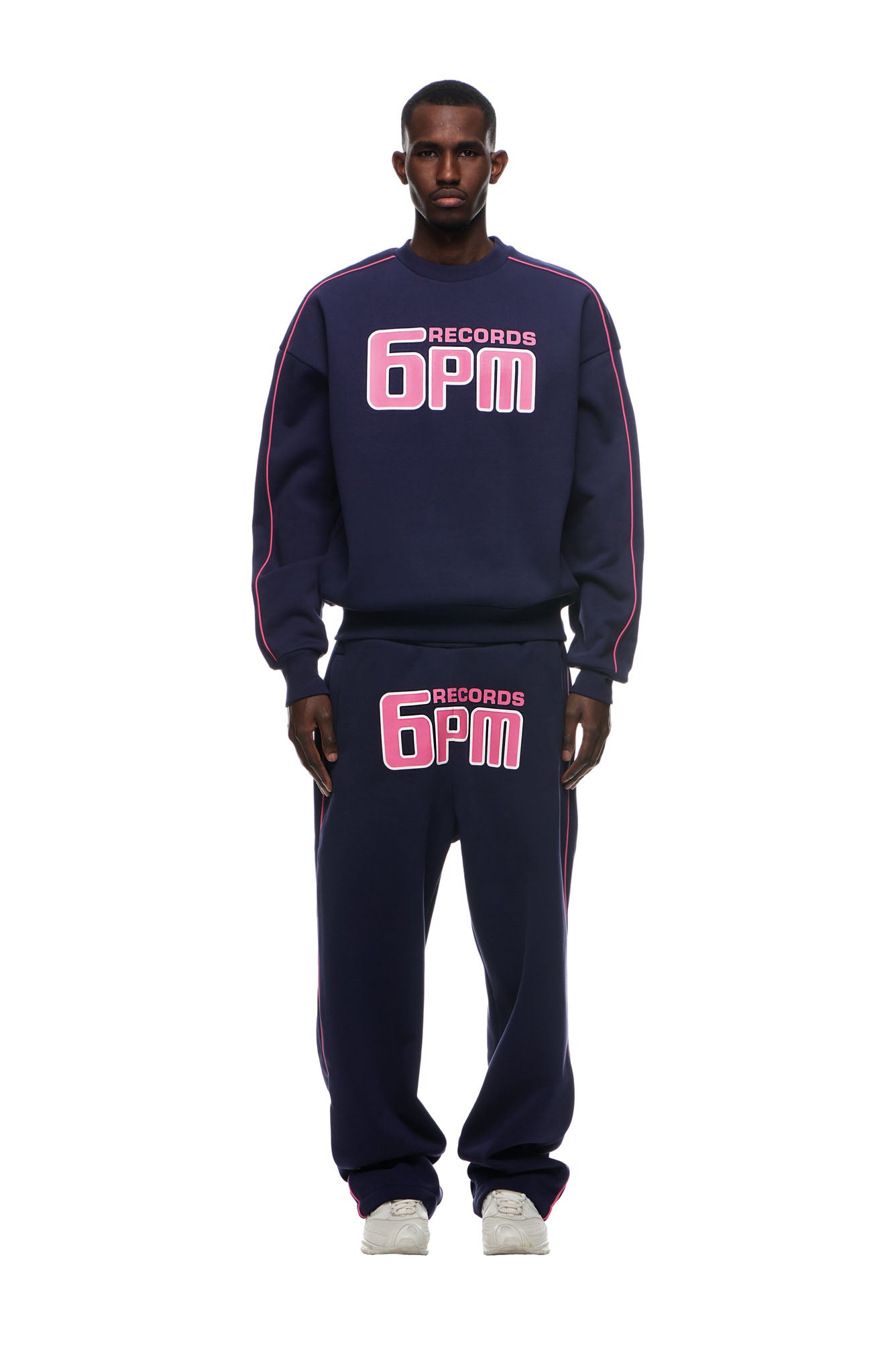 A man wearing the 6PM RECORDS CREWNECK DEEP BLUE sweatshirt and matching sweatpants with pink logos, white sneakers, and pink trim stands against a white background.