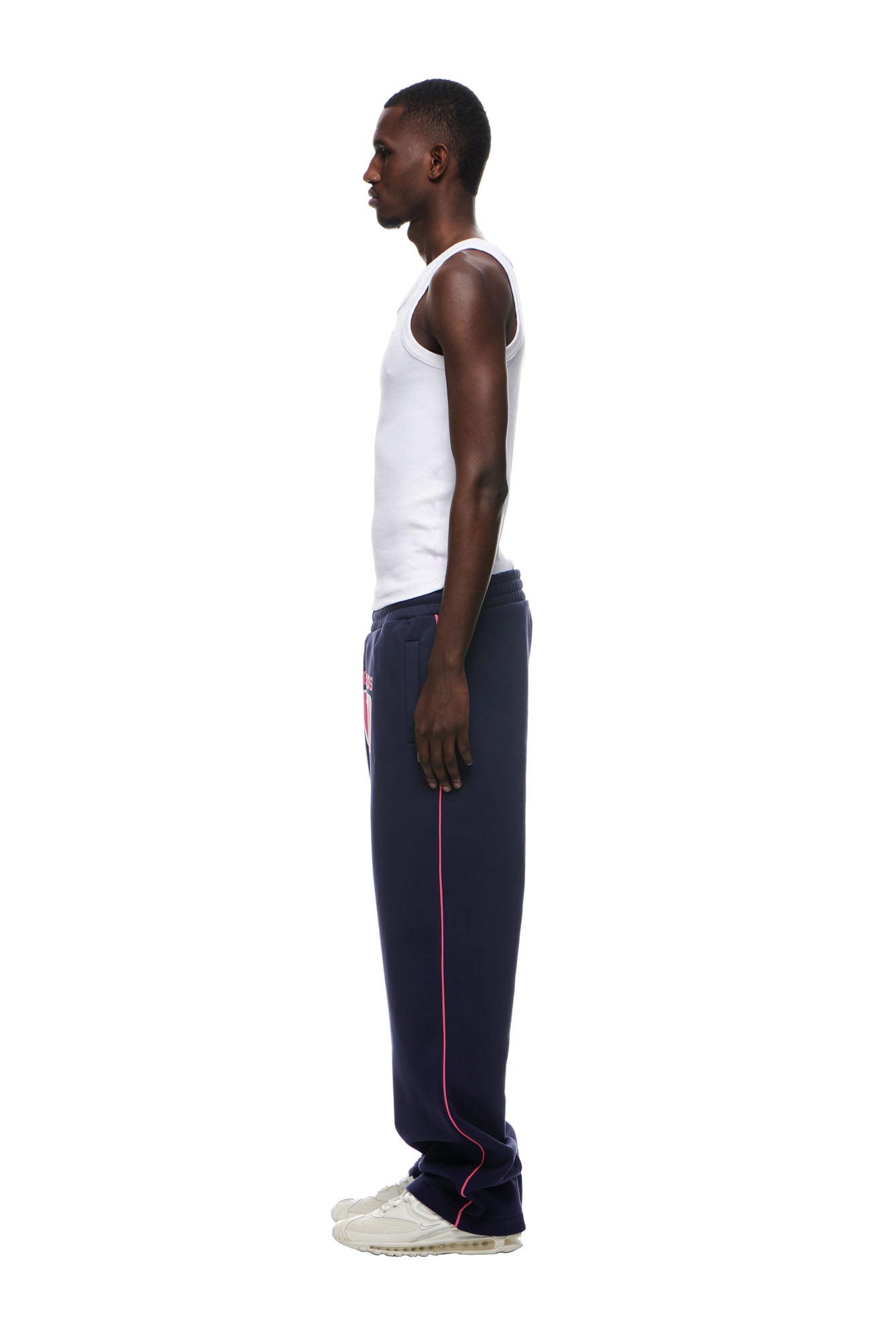 A man in profile faces left, wearing a white tank top, 6PM RECORDS OPEN LEG JOGGER DEEP BLUE sweatpants with pink piping, and white sneakers against a plain white background.