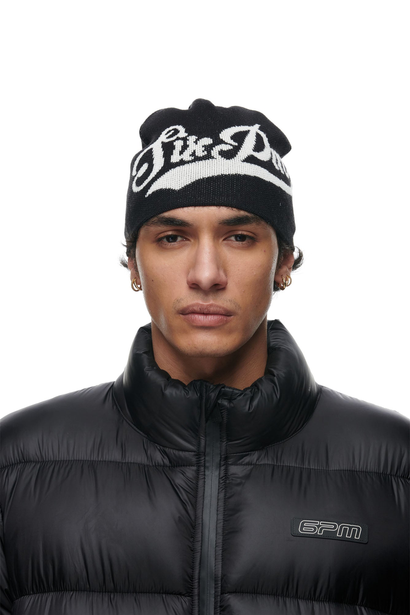 A young man with short hair and multiple earrings wears a 6PM SCRIPT LOGO BEANIE BLACK and a black puffer jacket, looking directly at the camera with a neutral expression.