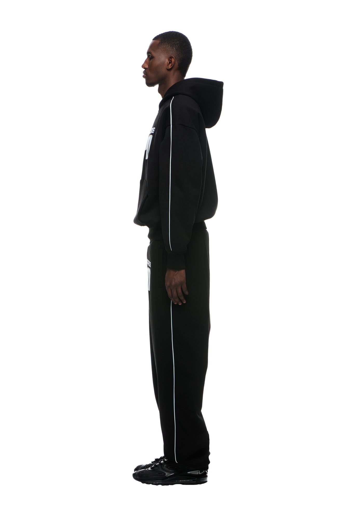 A person stands in profile against a white background, wearing the 6PM RECORDS HOODIE BLACK with the hood up, paired with black pants featuring white piping and black sneakers.