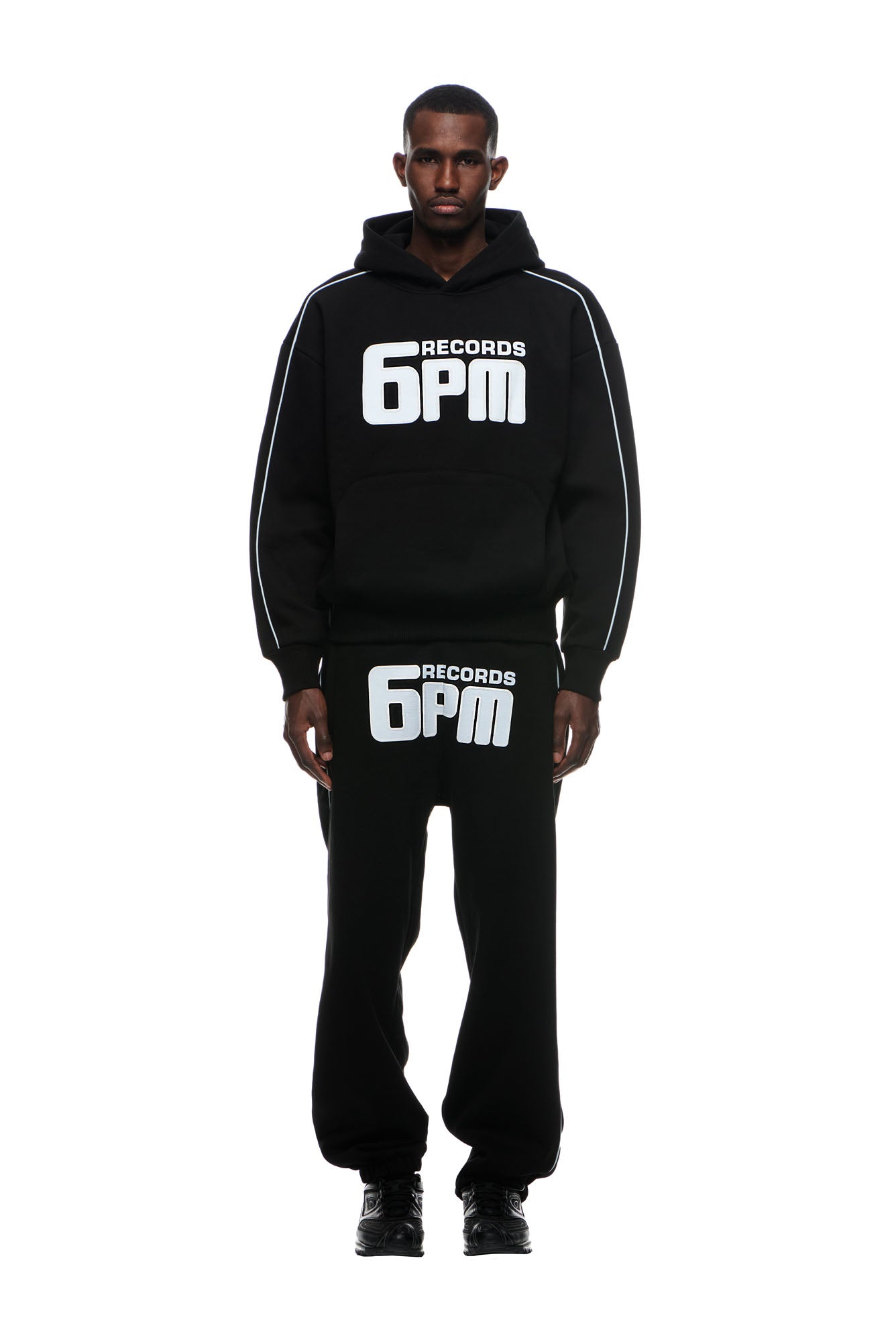 A man faces forward against a white background wearing the 6PM RECORDS HOODIE BLACK, featuring bold white "6PM RECORDS" lettering on the chest, paired with matching black sweatpants. He has a neutral expression.