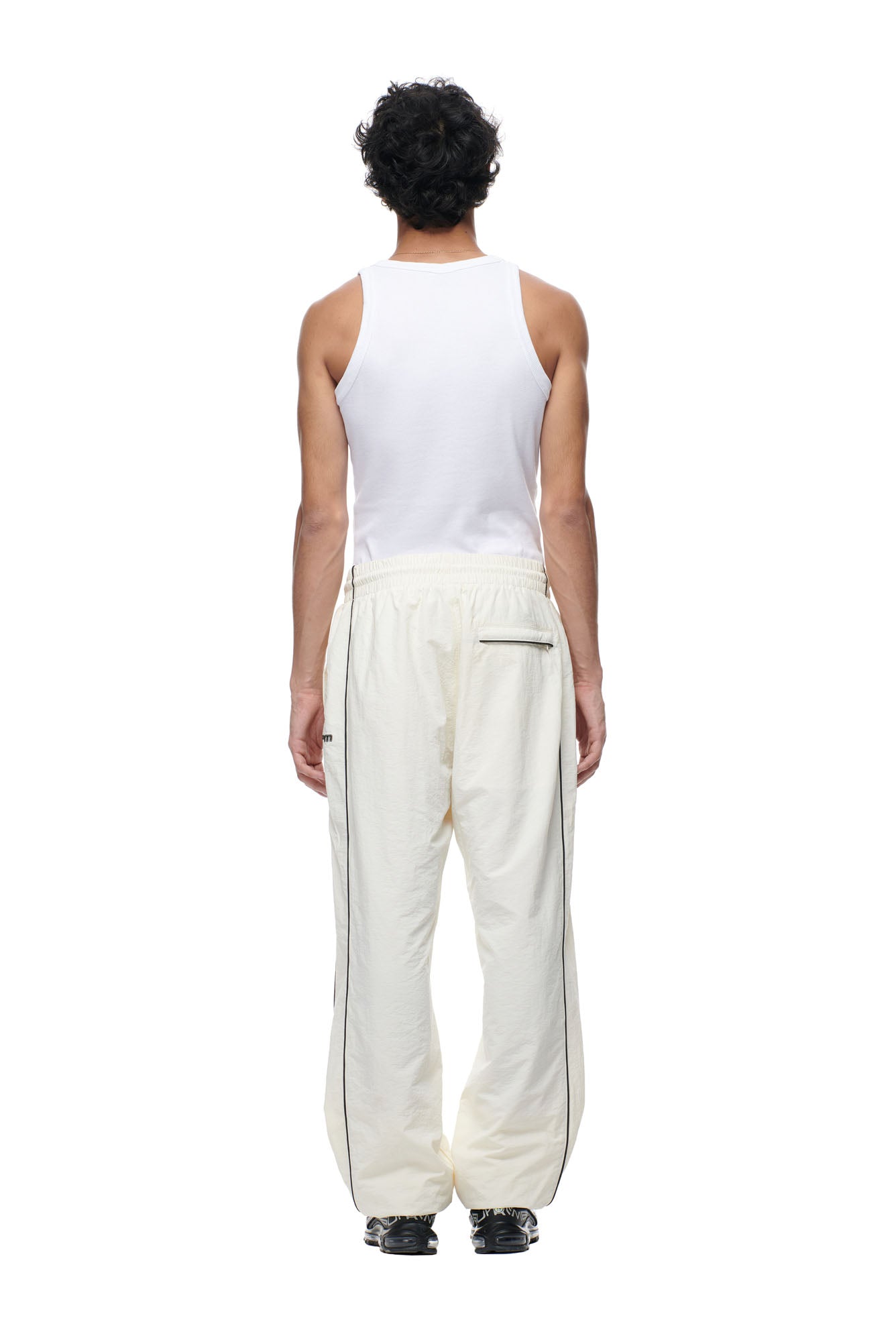 A person with short, curly hair is seen from behind wearing a white tank top, black shoes, and 6PM BF NYLON TRACKPANTS CREAM, featuring a back zipper pocket, standing against a plain white background.