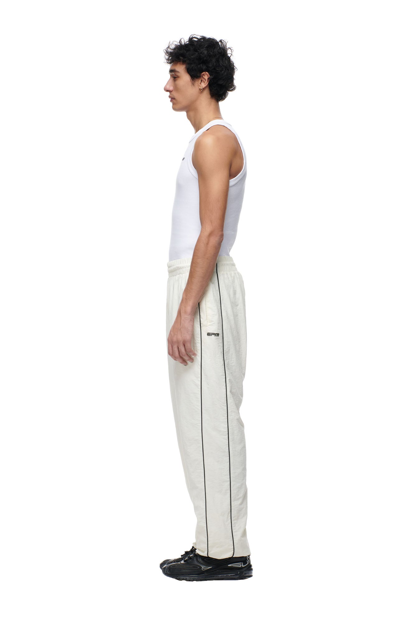 A man with curly dark hair stands in profile, wearing 6PM's BF NYLON TRACKPANTS CREAM, a white tank top, and black sneakers against a plain white background.