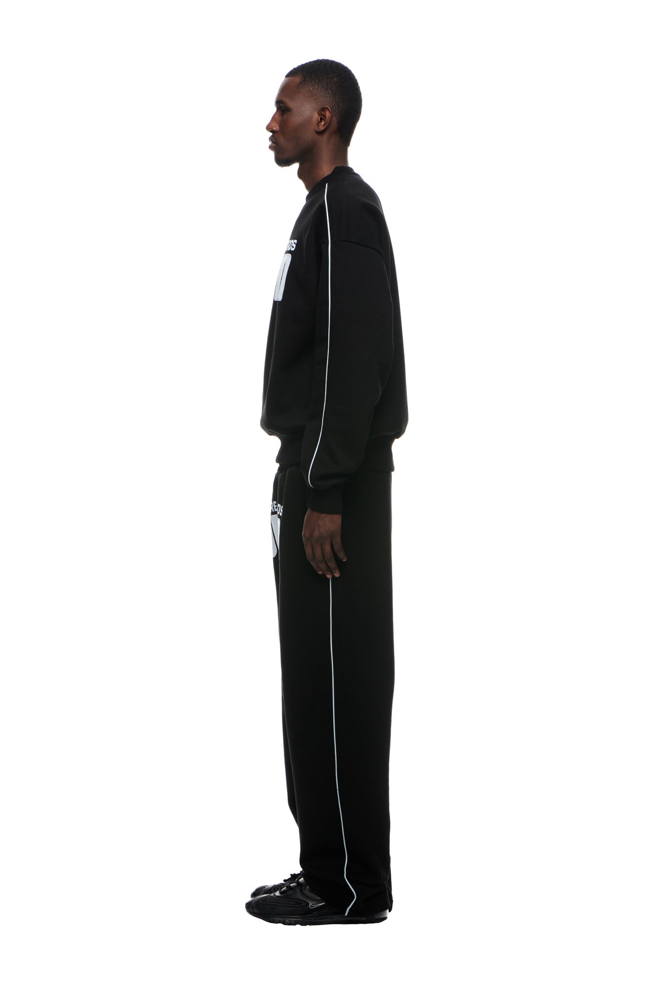 A man stands in profile against a white background, wearing the 6PM RECORDS CREWNECK BLACK sweatshirt with matching black pants featuring white piping and text, paired with black sneakers.