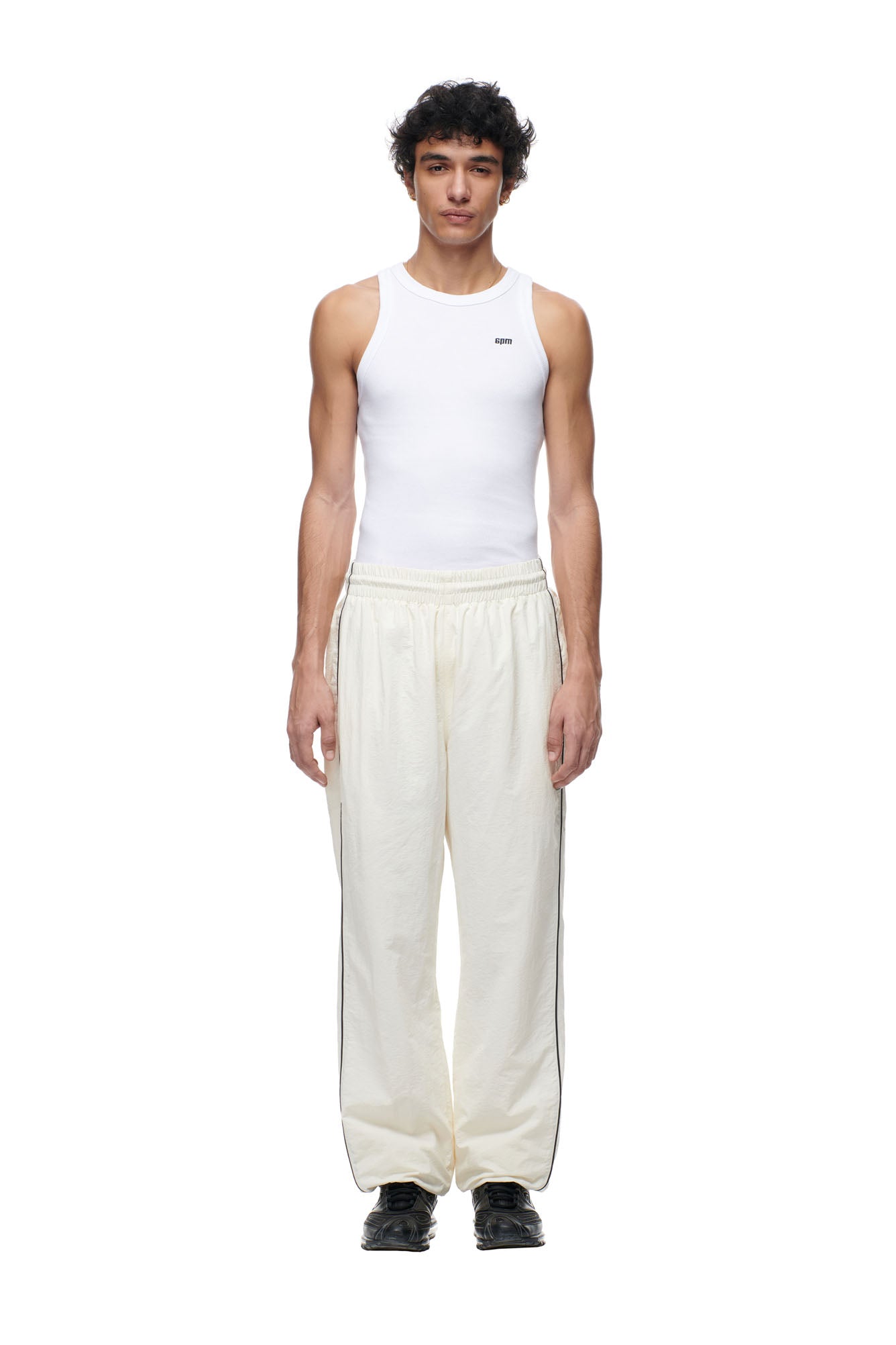 A young man faces forward in a fitted white tank top, BF NYLON TRACKPANTS CREAM by 6PM, and black athletic shoes against a plain white background.