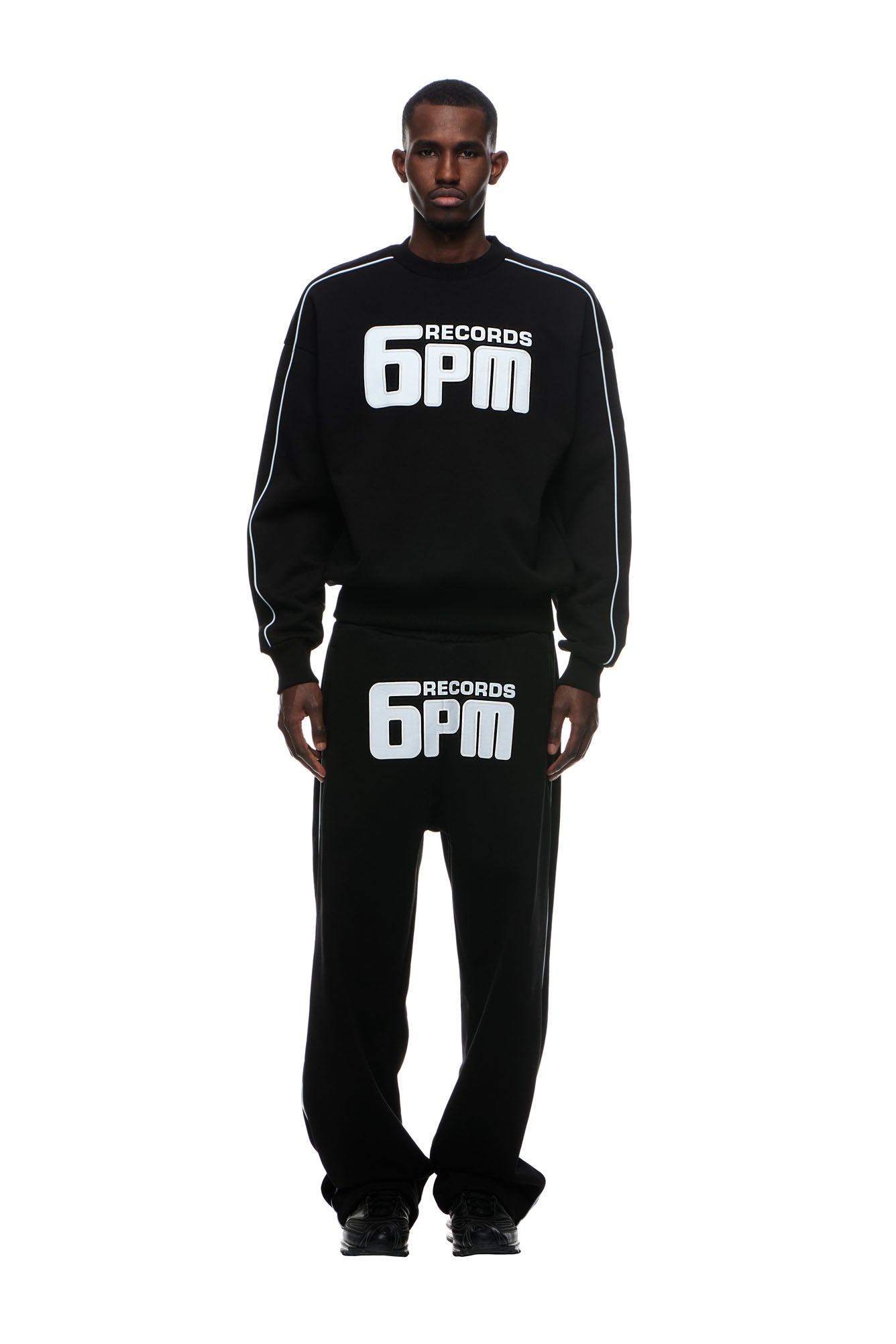 A man stands facing forward in the 6PM RECORDS CREWNECK BLACK sweatshirt and matching pants, both featuring bold white "6PM RECORDS" prints. He pairs the outfit with black shoes against a white background.