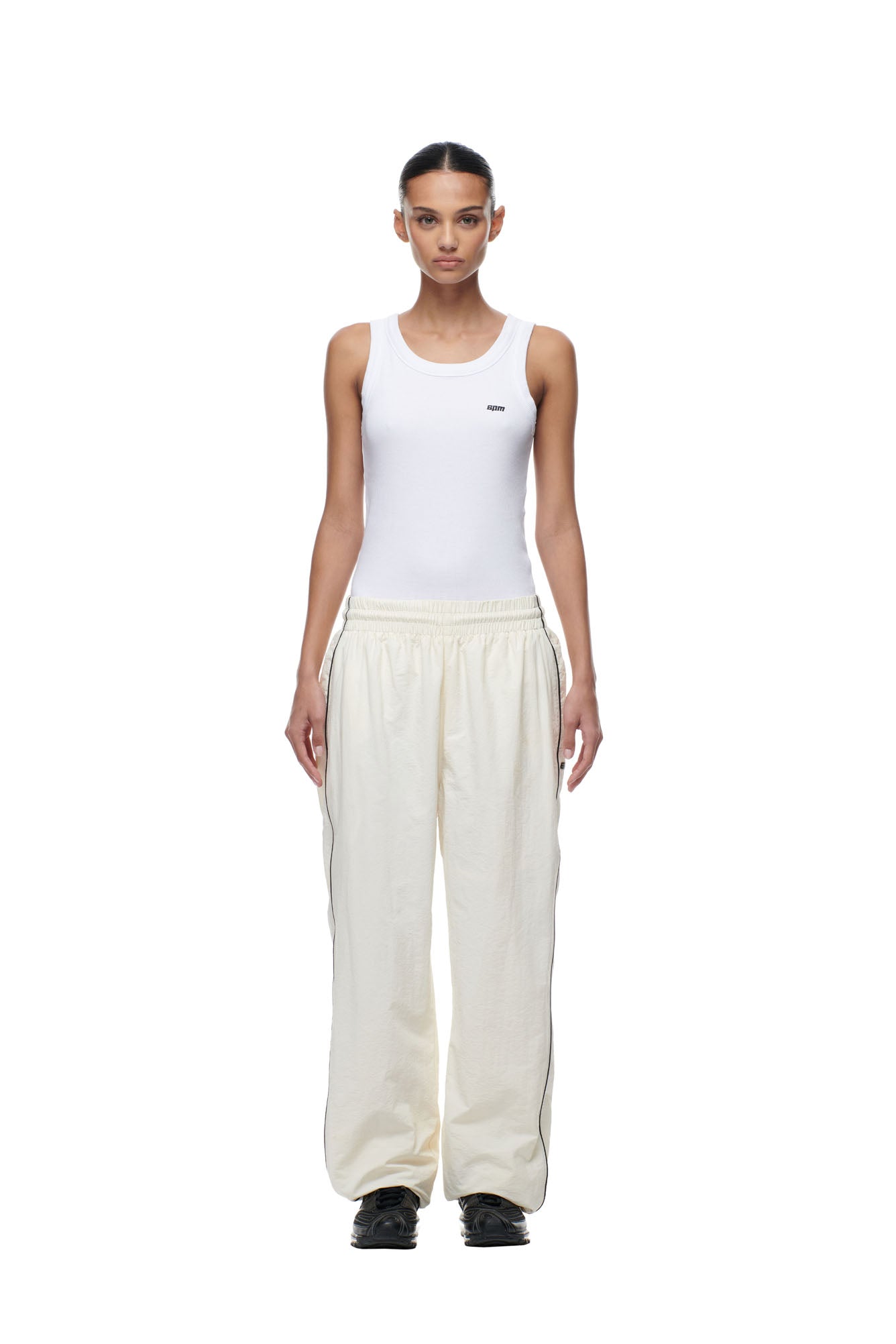 A woman stands facing forward, wearing the 6PM BF NYLON TRACKPANTS CREAM and a fitted white tank top with black shoes, against a plain white background.