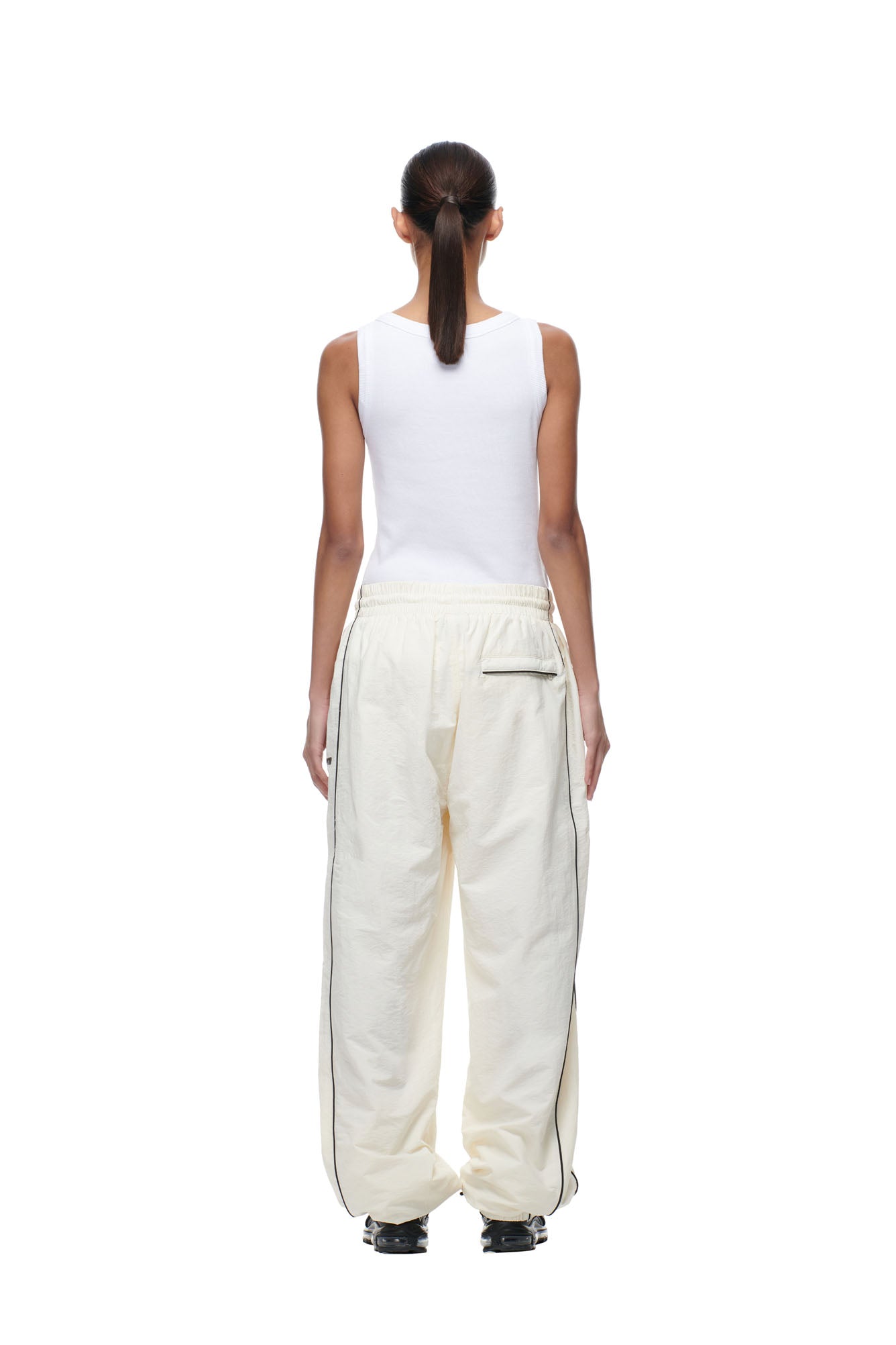 A person with long hair in a low ponytail, wearing a white sleeveless tank top and 6PM's BF NYLON TRACKPANTS CREAM, stands facing away against a plain white background.
