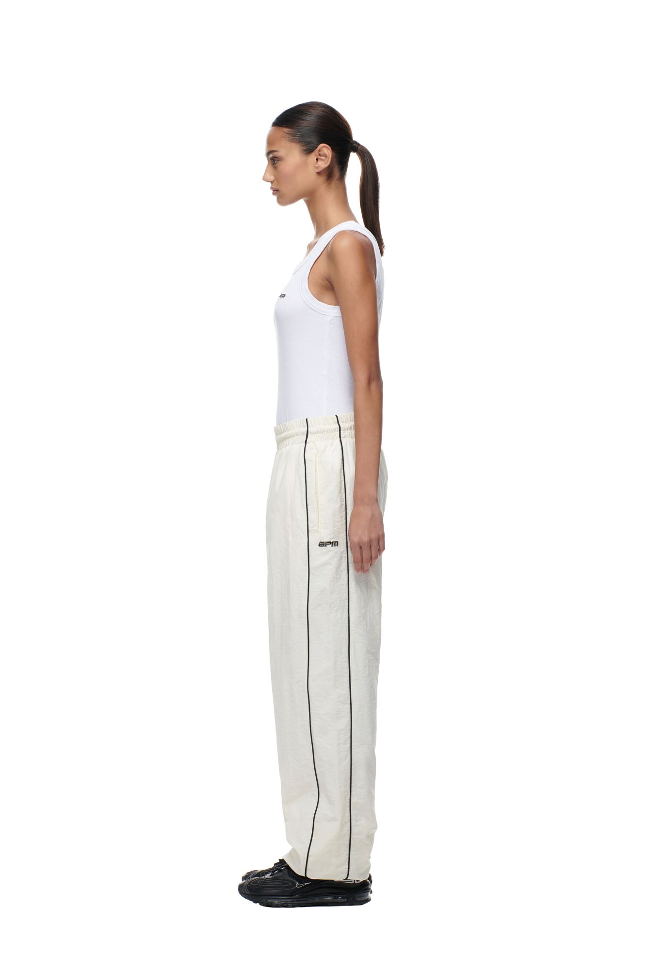 A woman stands in profile against a white background, wearing a white sleeveless top, 6PM's BF NYLON TRACKPANTS CREAM, and black shoes. Her hair is tied back in a low ponytail.