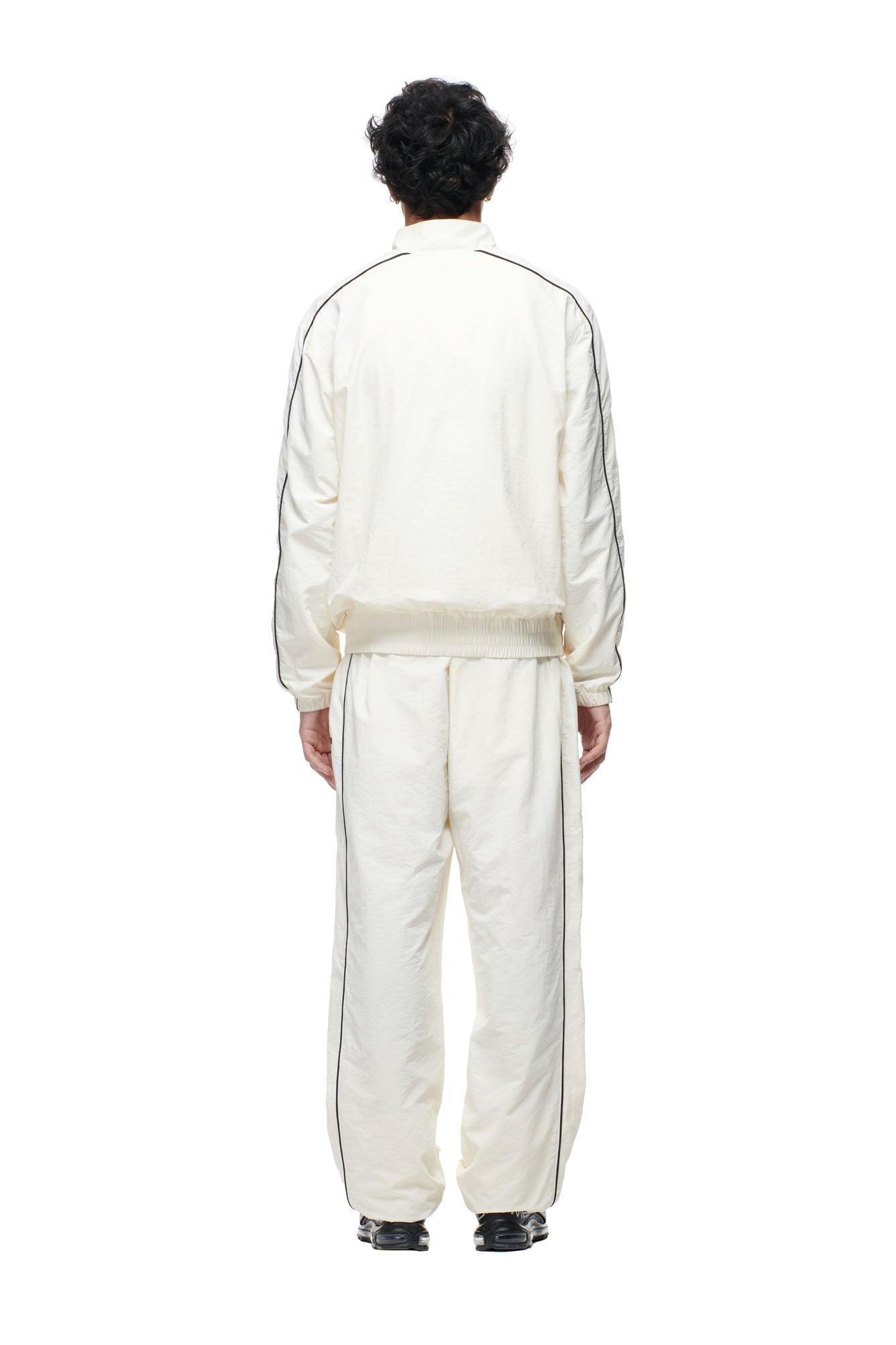 A person with short dark hair stands facing away, wearing the 6PM BF NYLON TRACKJACKET CREAM, featuring black stripes, against a plain white background.