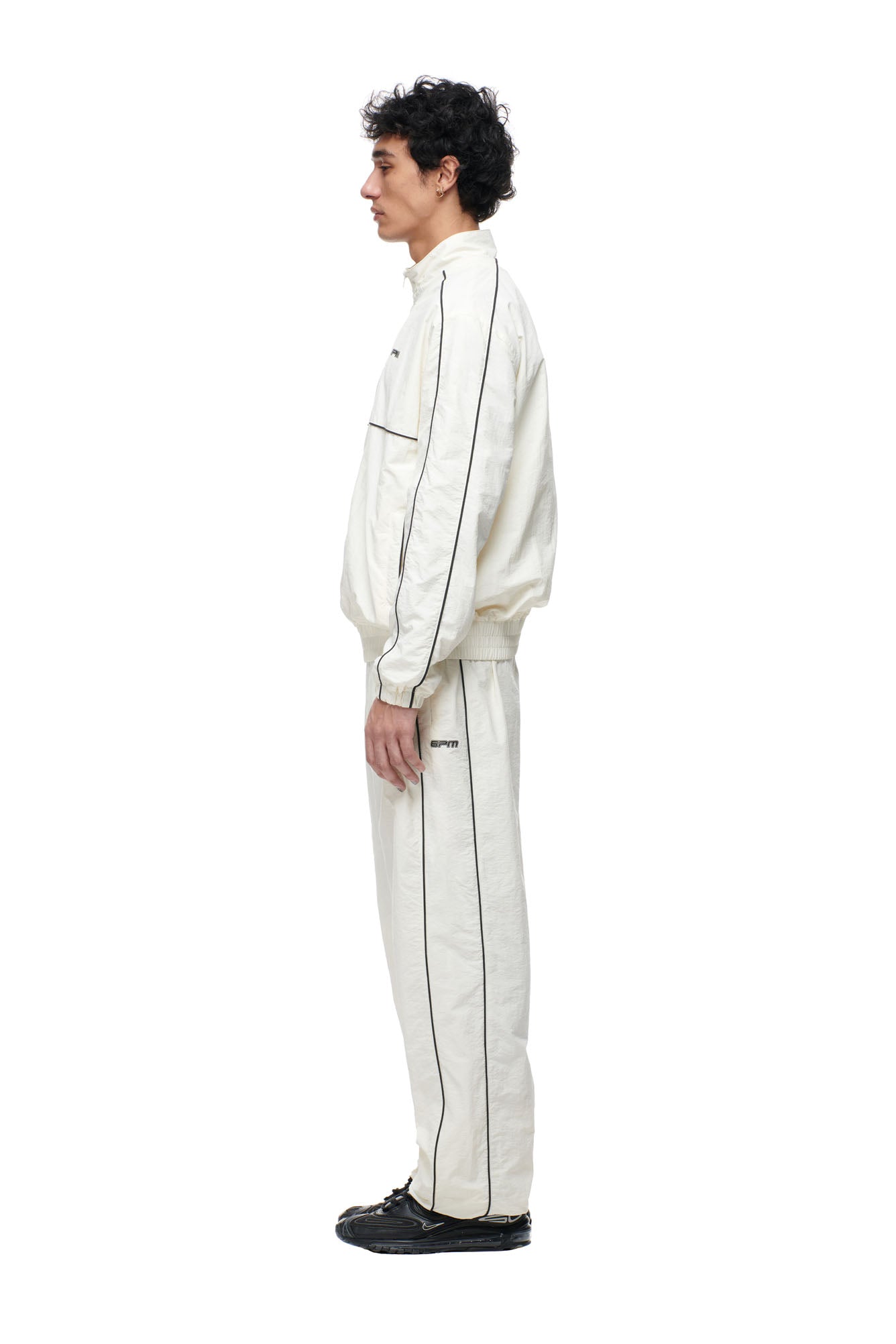 A man stands in profile against a white background, wearing the 6PM BF NYLON TRACKJACKET in cream with black piping and black shoes.