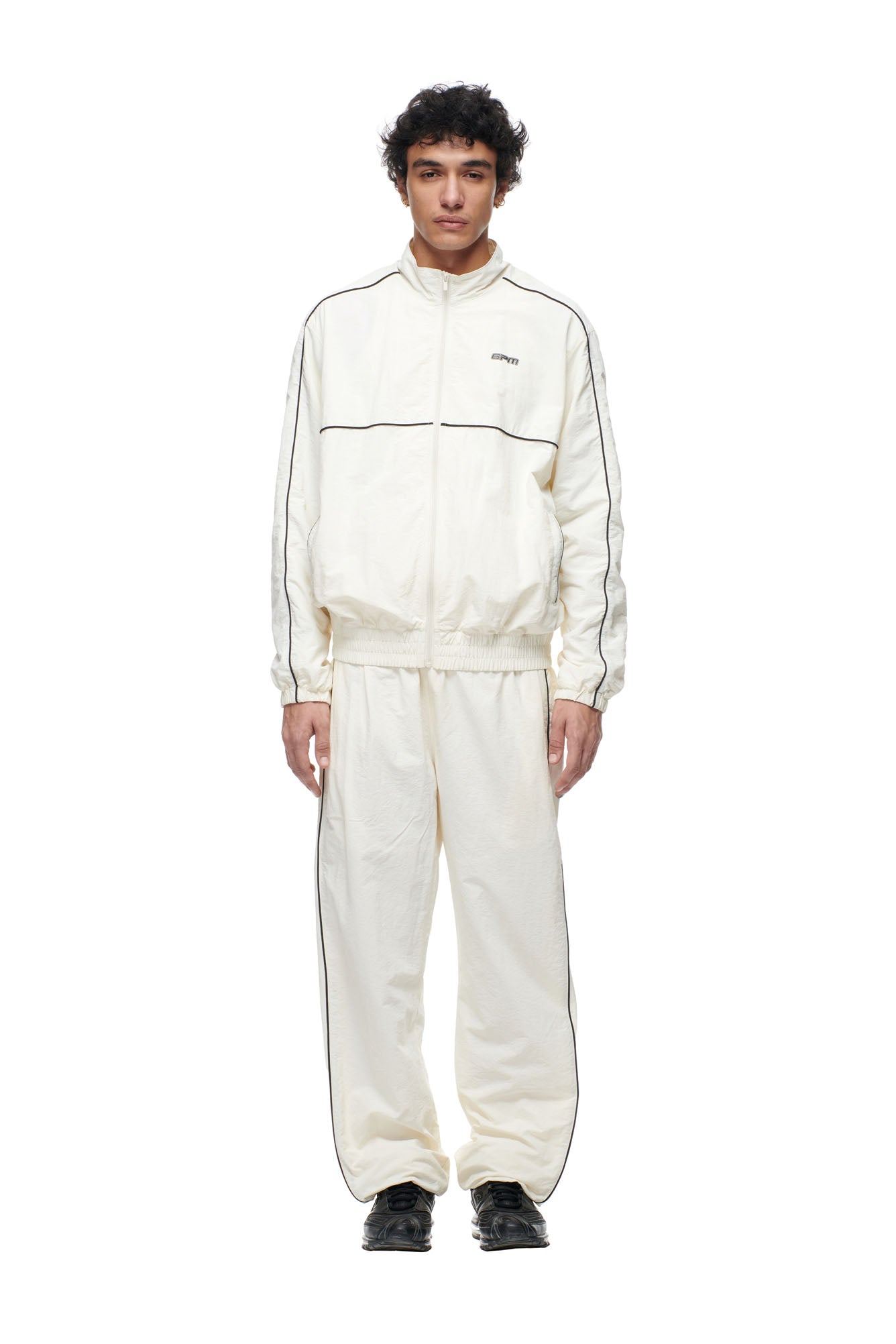 A person faces forward in a loose-fitting 6PM BF NYLON TRACKJACKET CREAM with black piping and matching pants, featuring a small chest logo. They wear black shoes against a plain white background.