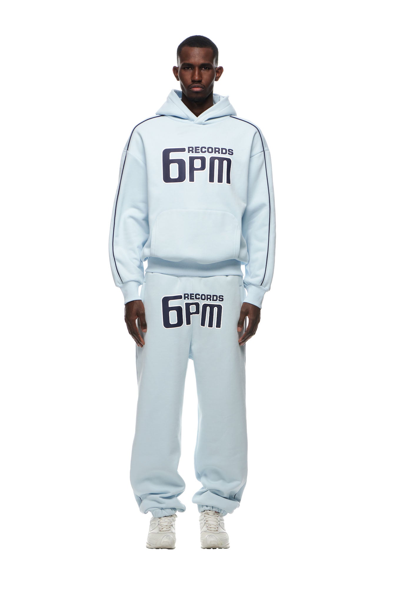 A man wears the 6PM RECORDS HOODIE ICE BLUE with matching sweatpants, both featuring bold black "6PM RECORDS" lettering. He pairs the set with white sneakers, standing against a plain white background.