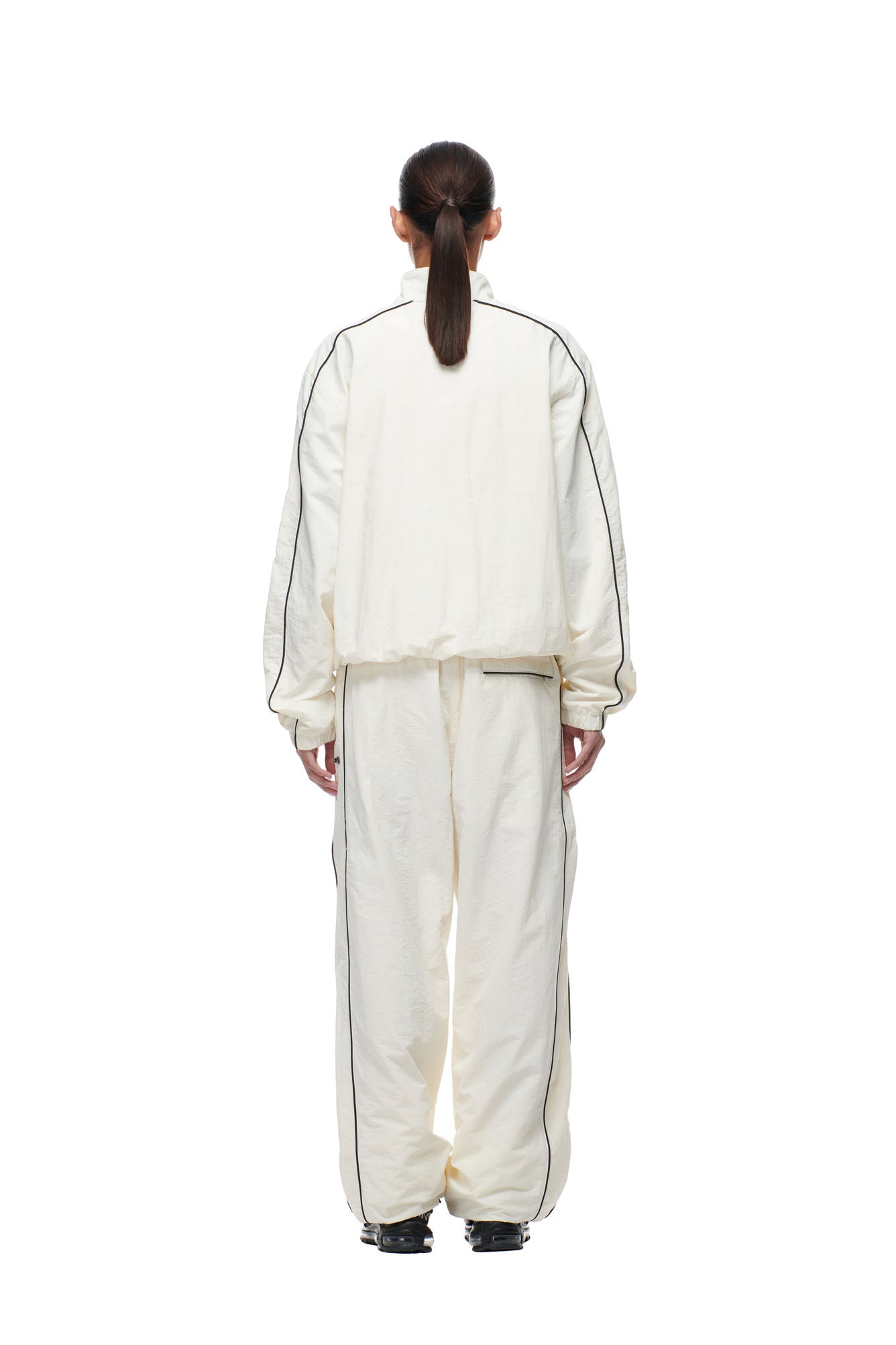 A person with dark hair in a low ponytail stands facing away, wearing the 6PM BF NYLON TRACKJACKET CREAM, a loose-fit off-white tracksuit with black piping, paired with black shoes, on a white background.