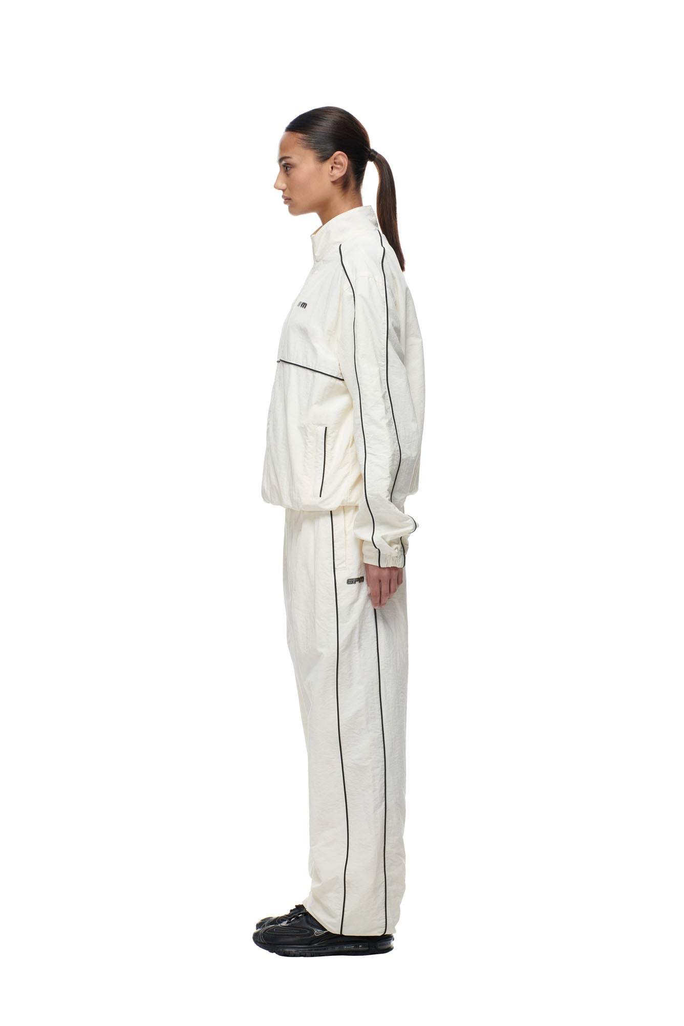 A woman with dark hair tied back stands in profile, wearing the 6PM BF NYLON TRACKJACKET CREAM, a loose-fitting off-white tracksuit with black piping, paired with black sneakers, set against a plain white background.