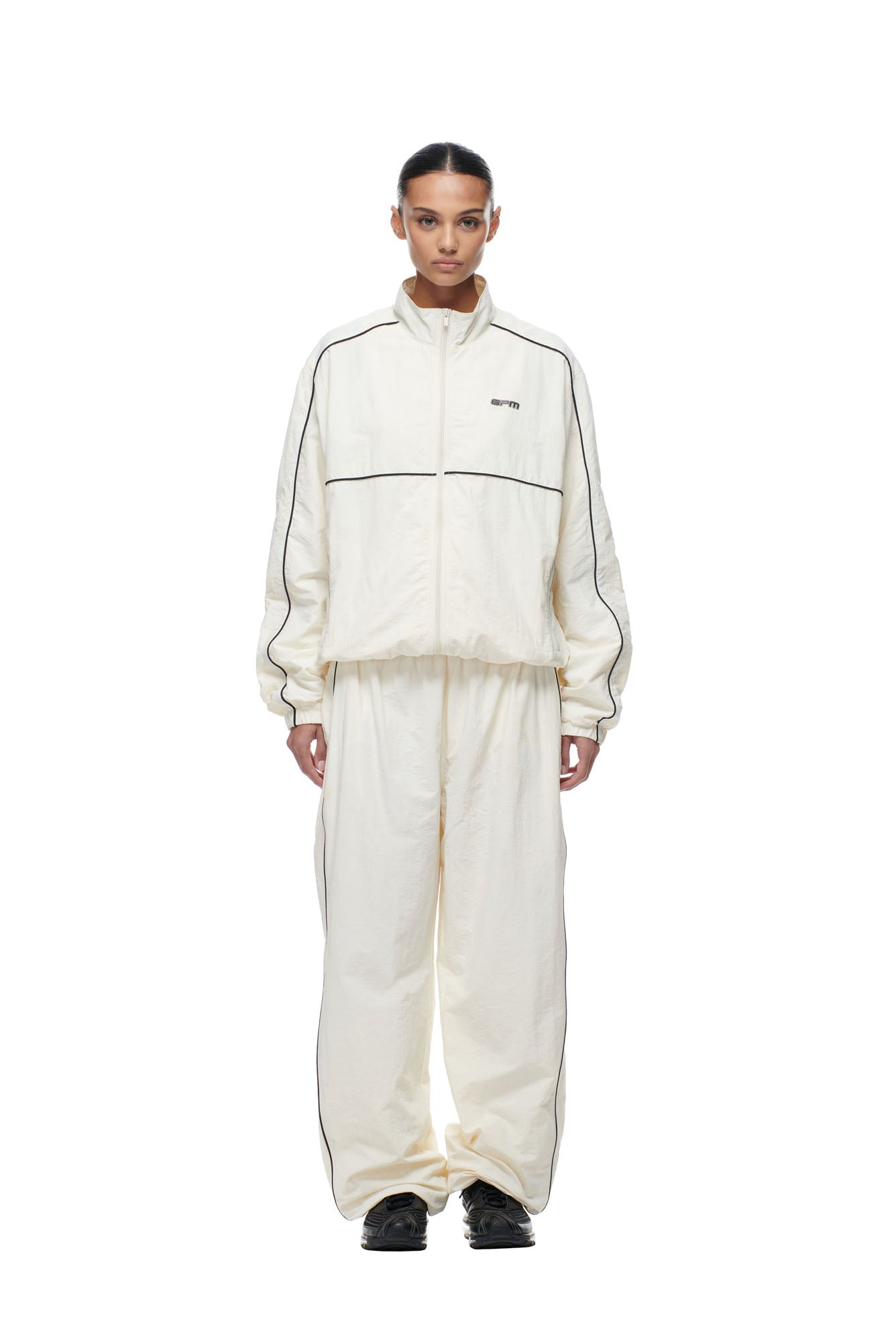 A person stands facing forward against a white background, wearing the 6PM BF NYLON TRACKJACKET CREAM—an off-white, loose-fitting track jacket with black piping, a zip front, and a small chest logo, paired with black shoes.