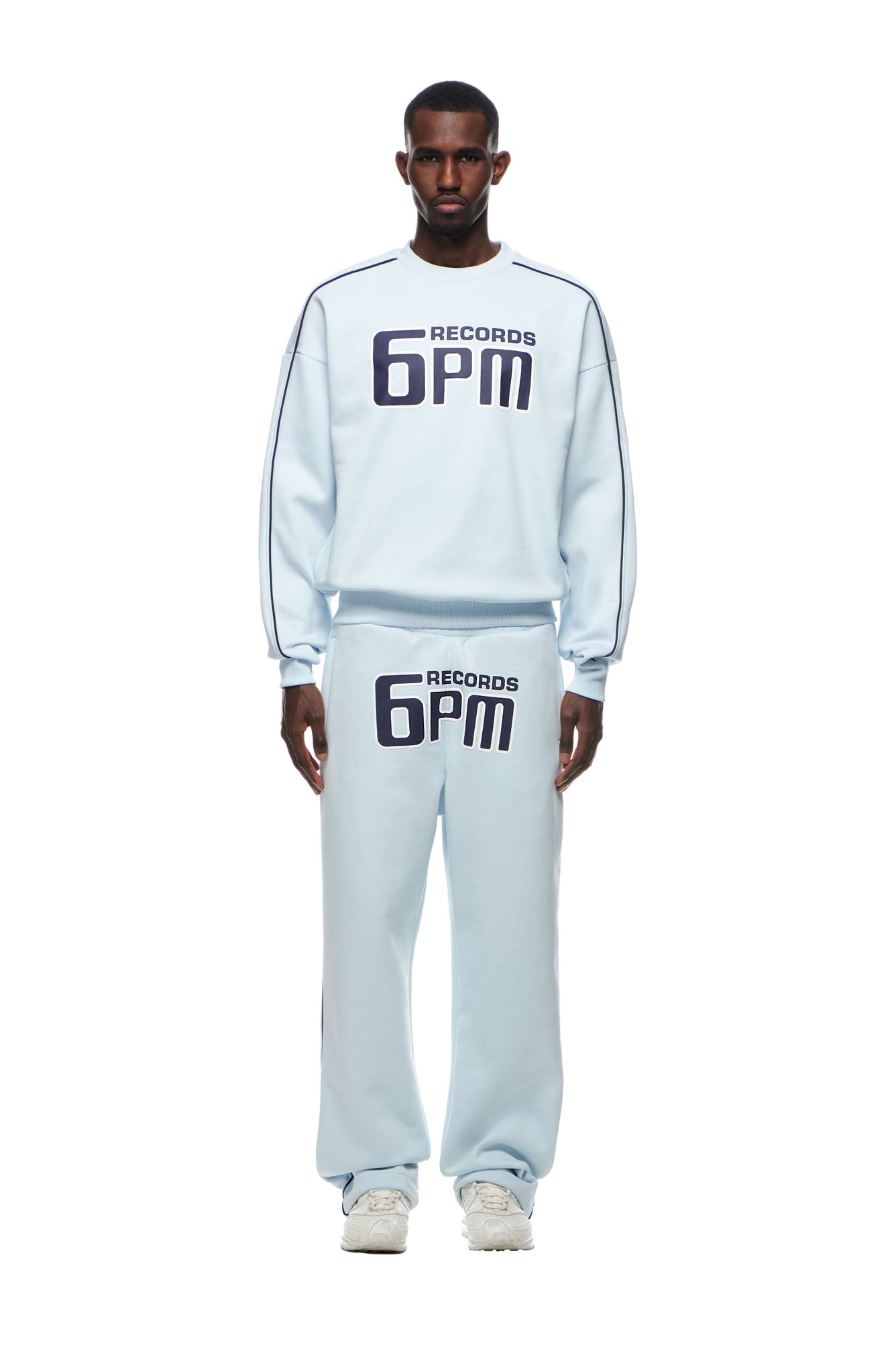 A man stands facing forward in the 6PM RECORDS CREWNECK ICE BLUE sweatshirt and matching sweatpants, both with bold black 6PM RECORDS lettering on the chest and thigh. He pairs the set with white sneakers against a plain white backdrop.