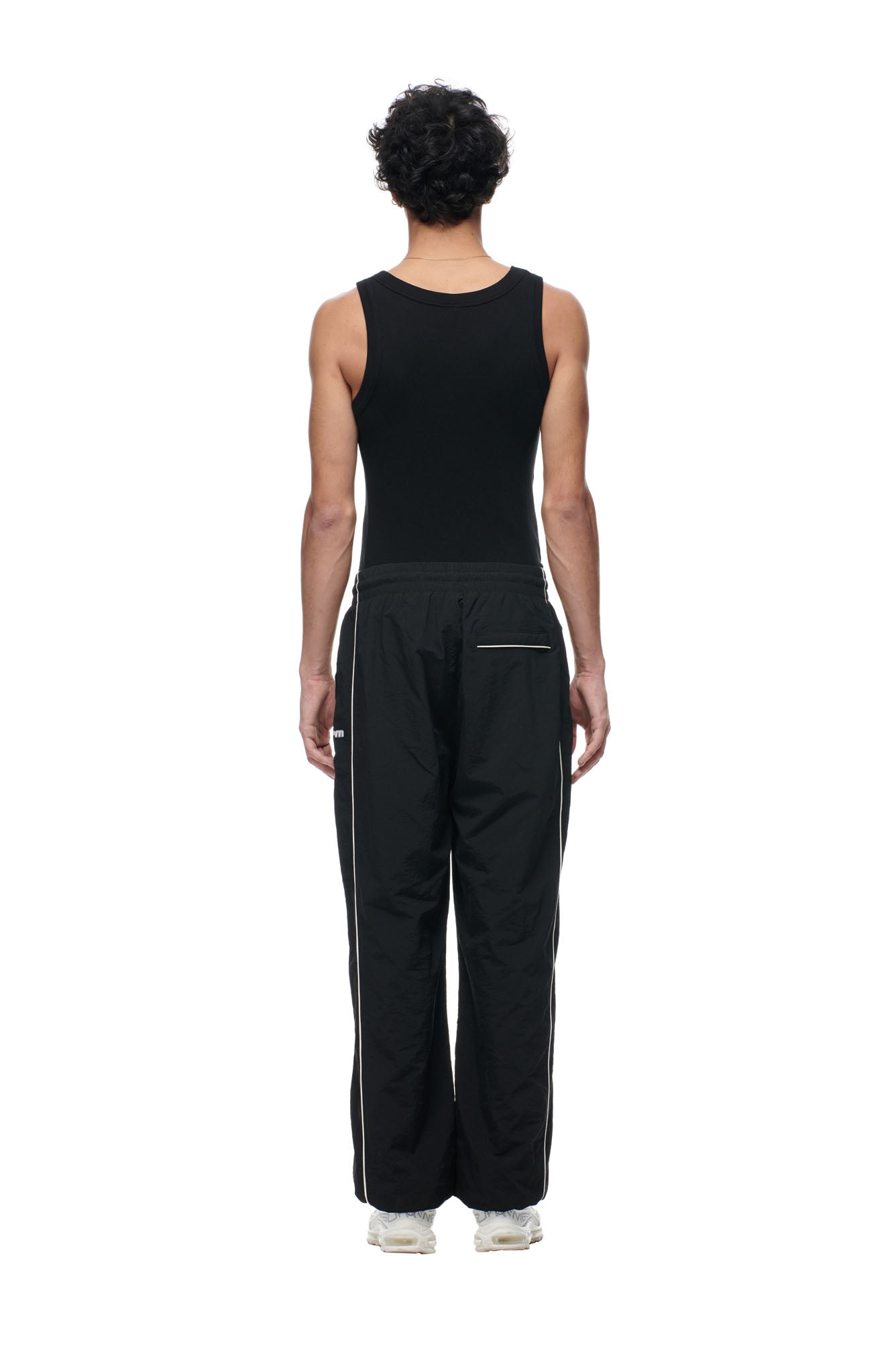 A person stands facing away from the camera, wearing 6PM BF NYLON TRACKPANTS BLACK, a black tank top, and white sneakers against a plain white background.