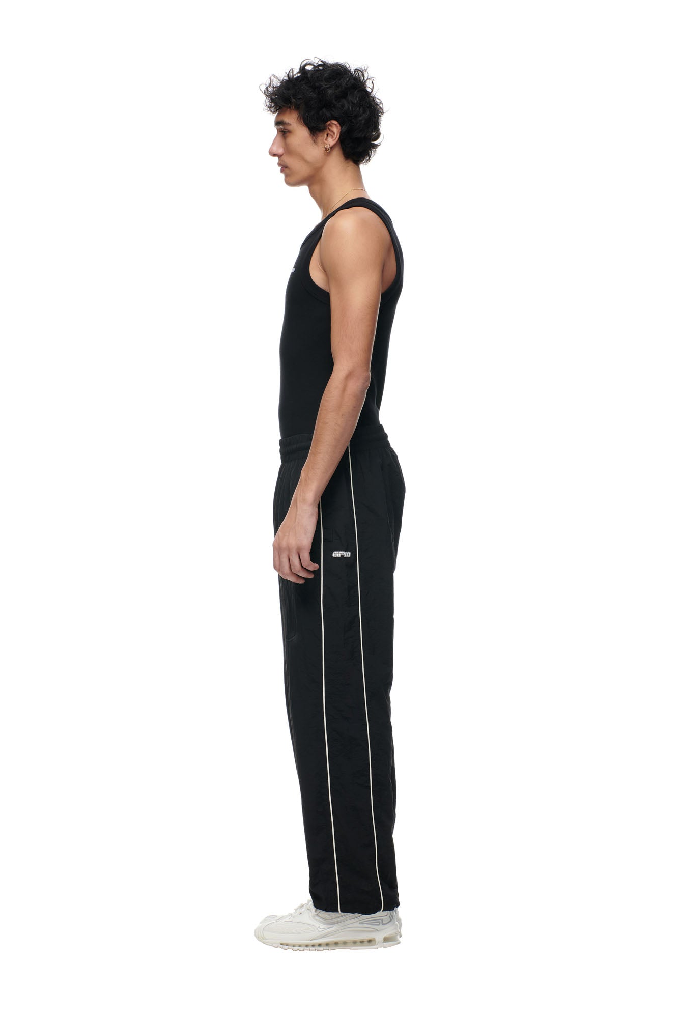 A man with curly hair stands in profile against a plain white background, wearing 6PM’s BF NYLON TRACKPANTS BLACK with white piping, a black sleeveless top, and white sneakers.