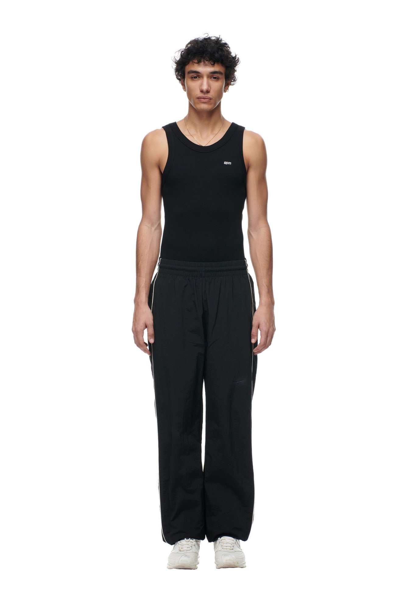 A young man stands facing forward in 6PM's BF NYLON TRACKPANTS BLACK, styled with white stripes, a black sleeveless tank top, and white sneakers against a plain white background.