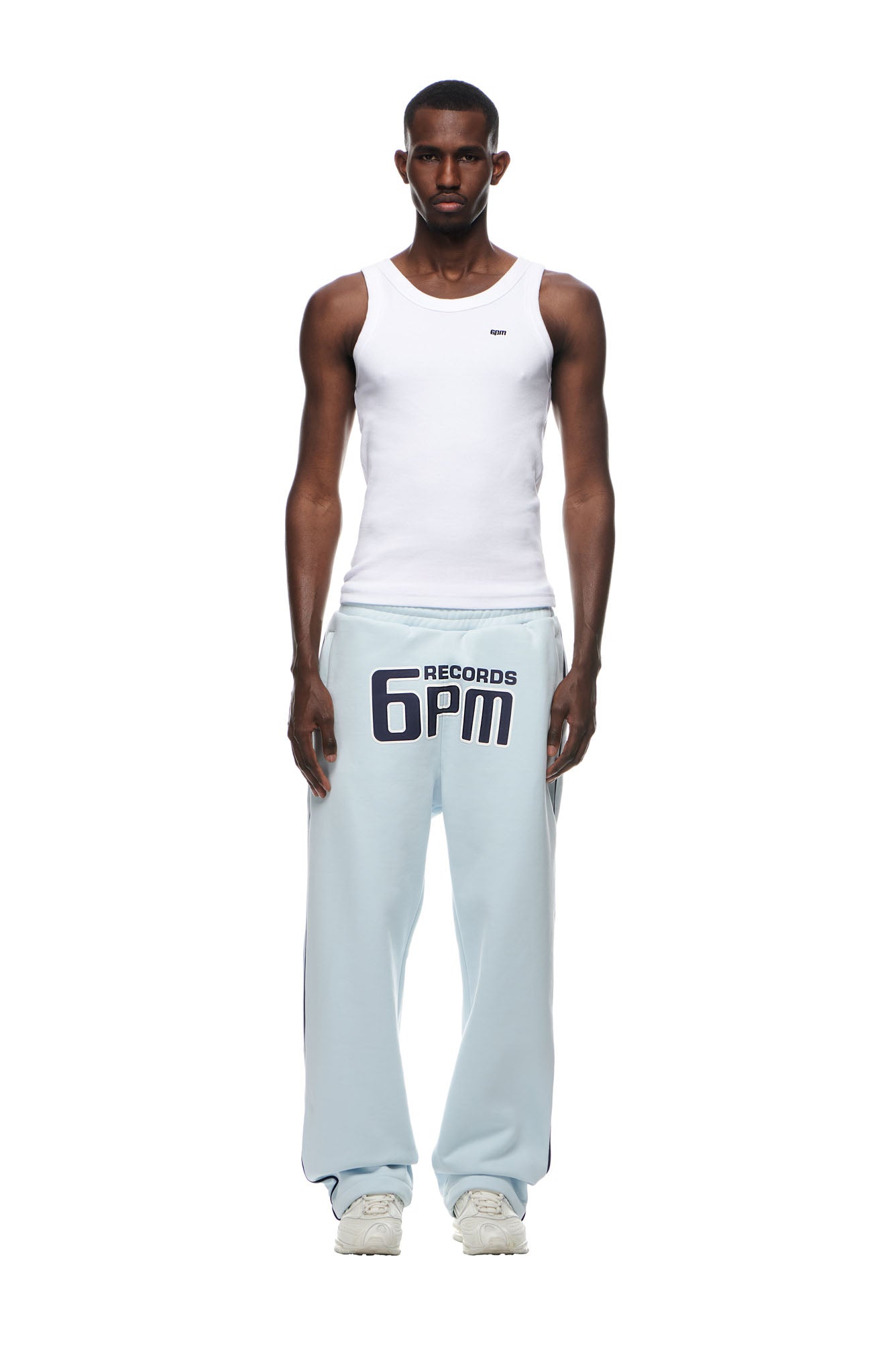 A man stands facing forward, wearing 6PM's RECORDS OPEN LEG JOGGER in Ice Blue with bold "6PM RECORDS" lettering, paired with a white tank top and white sneakers, against a plain white background.