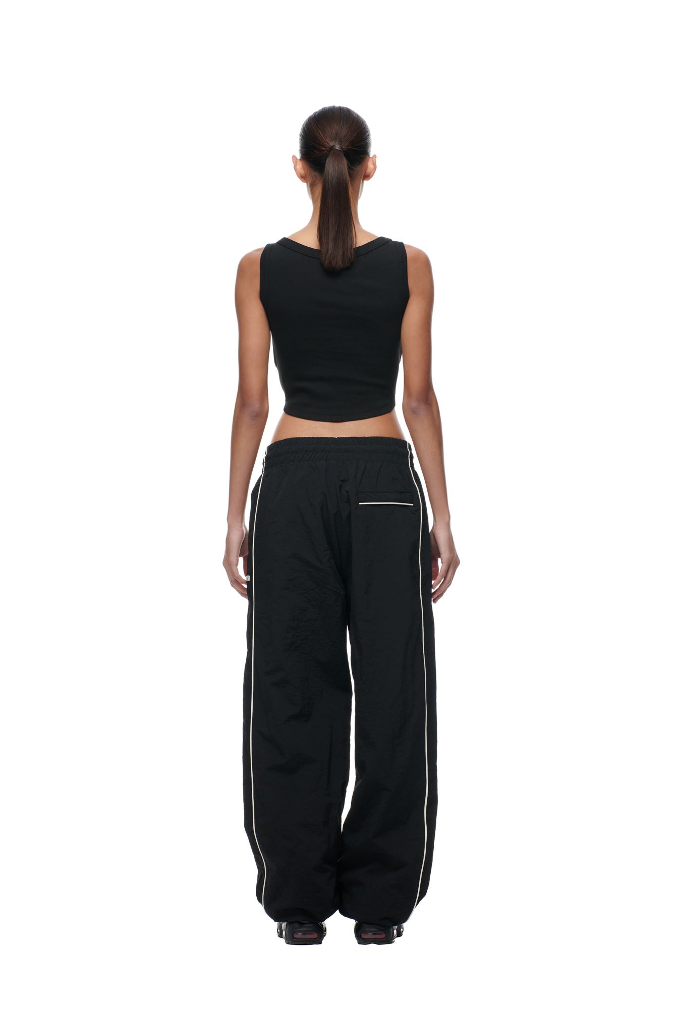 A woman faces away, wearing 6PM's BF NYLON TRACKPANTS BLACK—loose black pants with white side piping and a back zipper pocket—paired with a black sleeveless crop top, set against a white background.