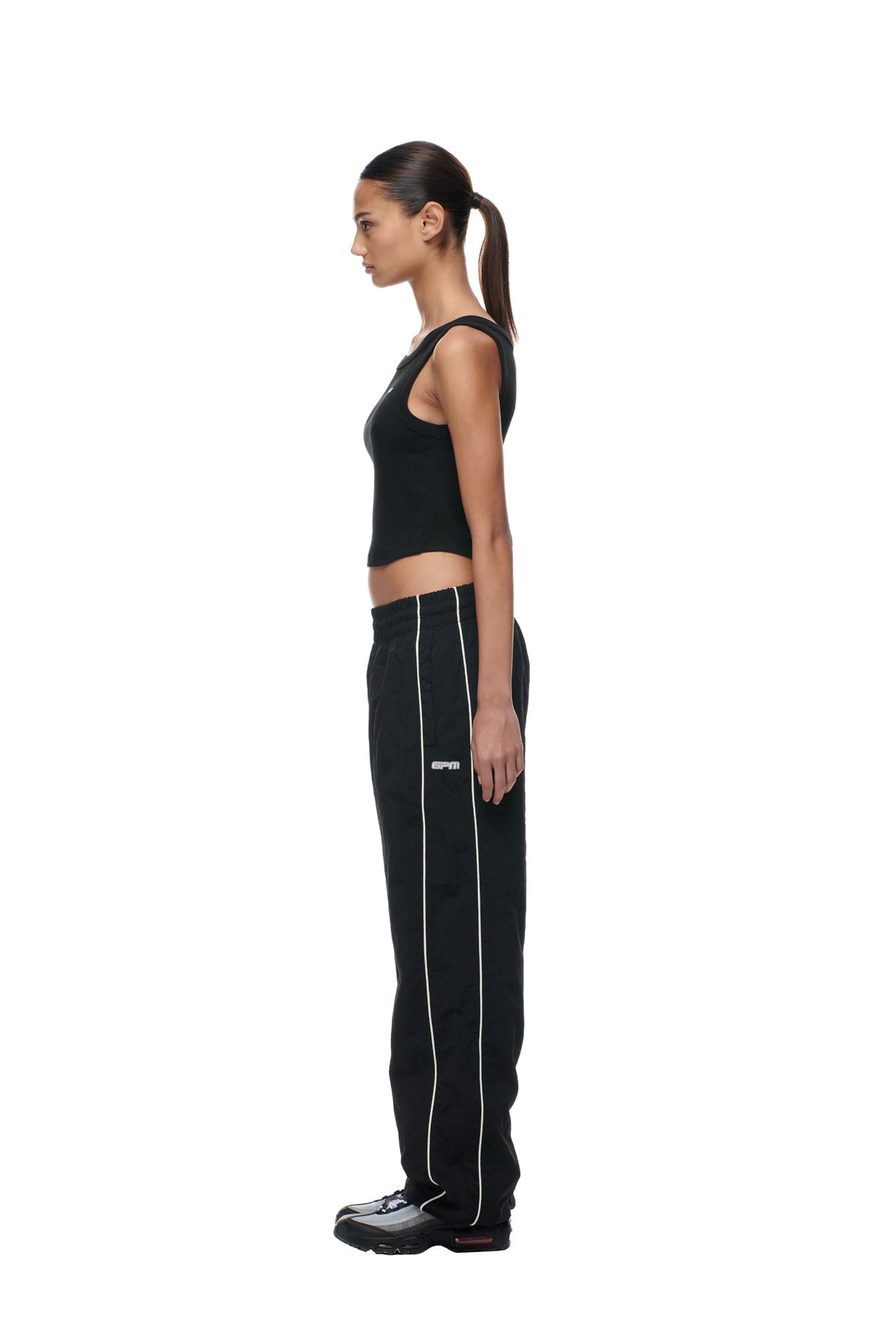 A woman stands in profile against a white background, wearing 6PM's BF NYLON TRACKPANTS BLACK with white piping, a black sleeveless top, black sneakers, and her hair tied back in a ponytail.