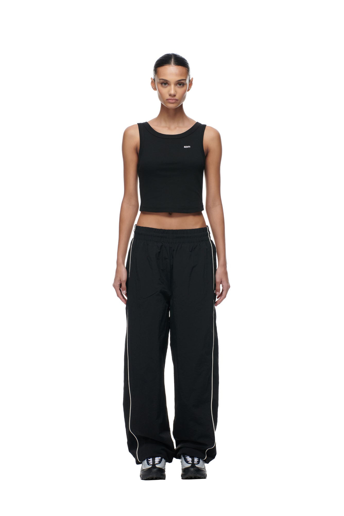 A person stands facing forward against a white background, wearing a black sleeveless crop top, 6PM BF NYLON TRACKPANTS BLACK with white piping, and black sneakers.