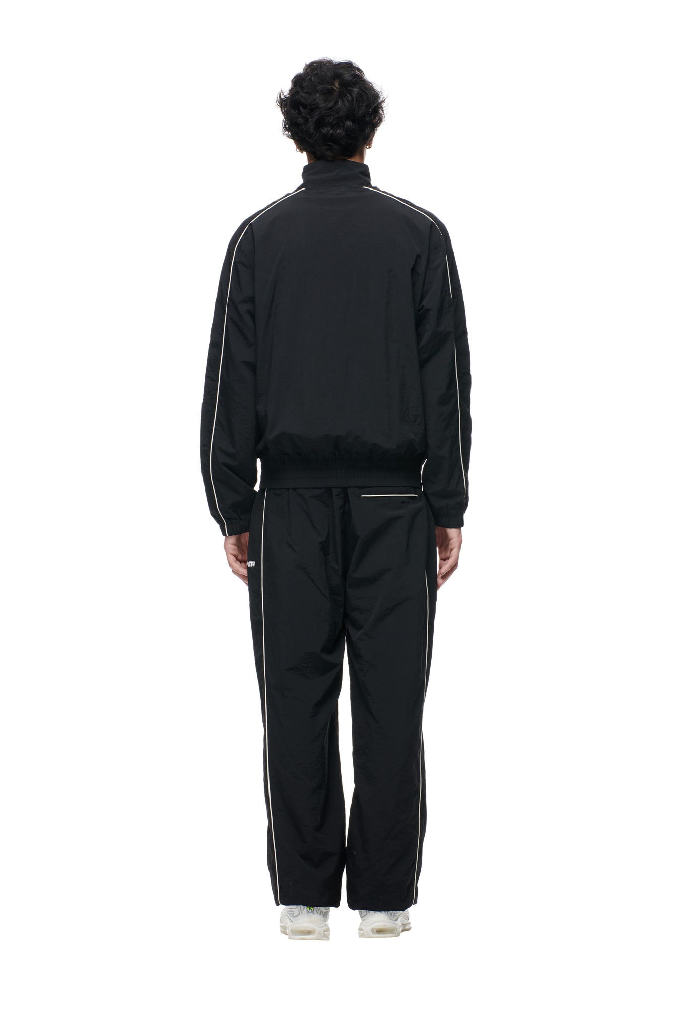 A person faces away from the camera, wearing the 6PM BF NYLON TRACKJACKET BLACK and white sneakers against a plain white background.