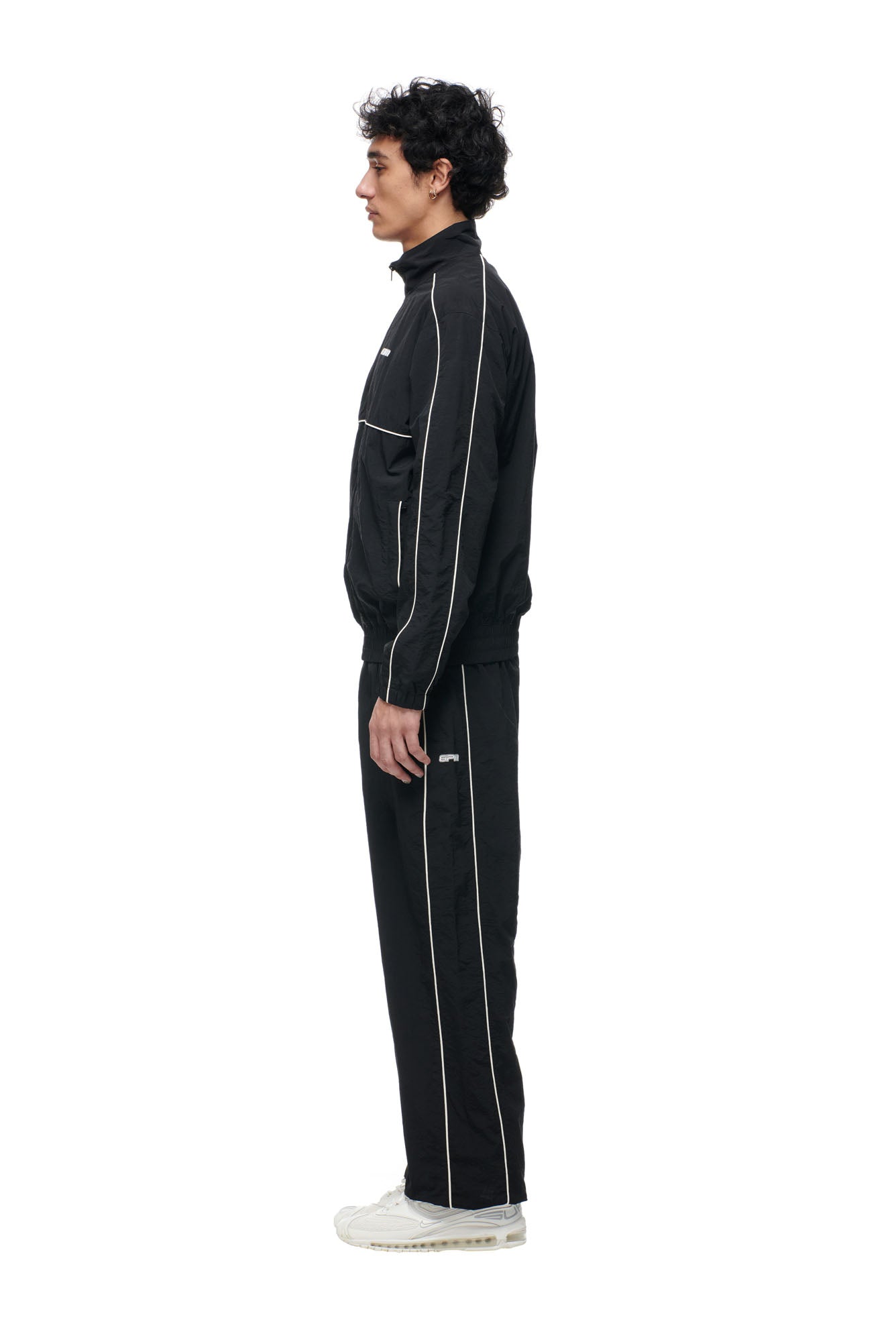 A person stands in profile against a white background, wearing the 6PM BF NYLON TRACKJACKET BLACK with white piping and white sneakers.
