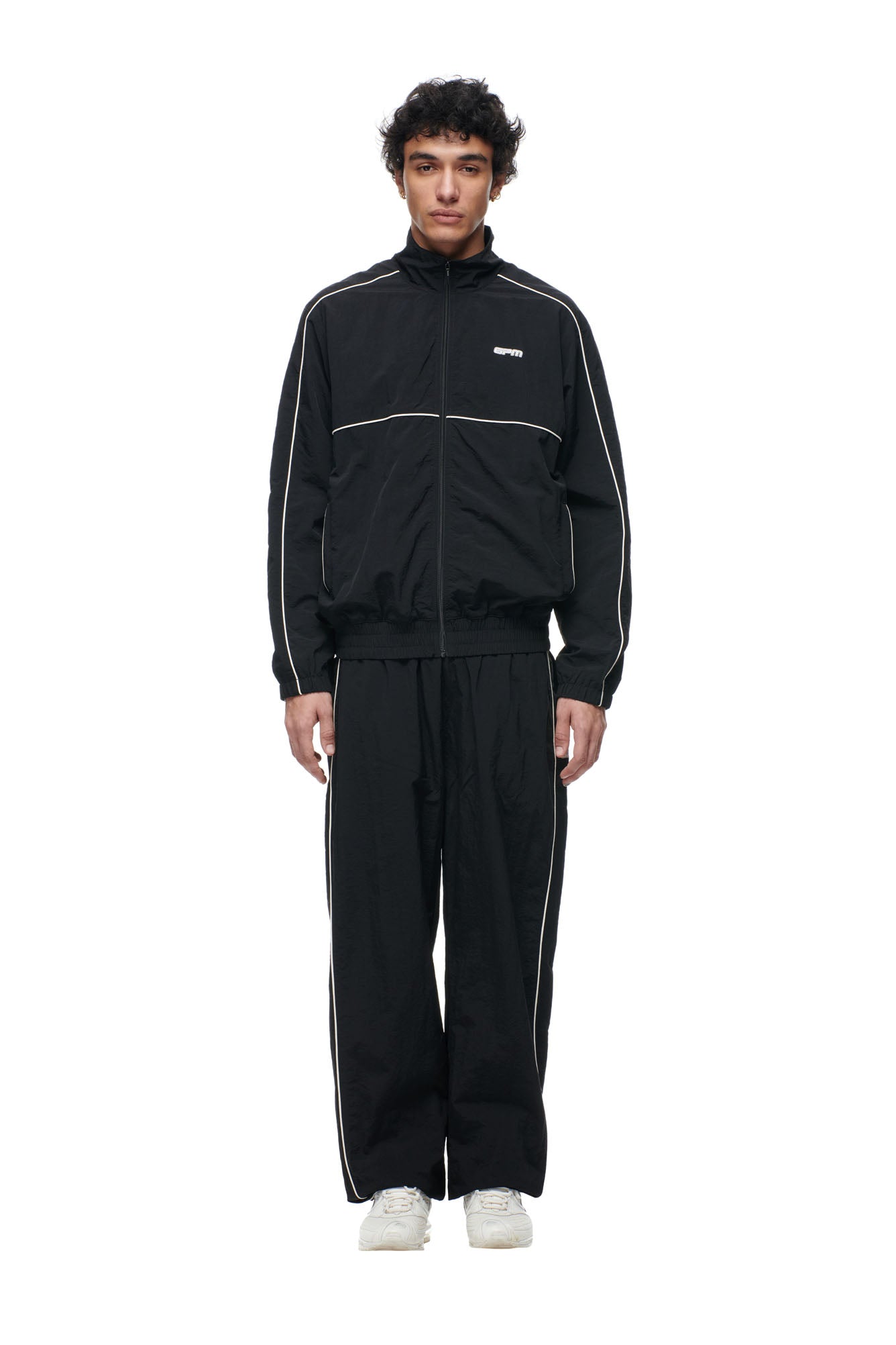 A person faces forward against a plain white background, wearing the 6PM BF NYLON TRACKJACKET BLACK, styled with white piping and paired with white sneakers.
