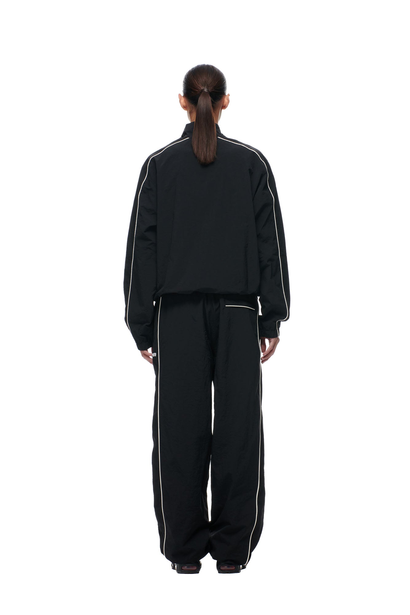A person with long dark hair in a ponytail stands facing away, wearing the 6PM BF NYLON TRACKJACKET BLACK, a loose-fit black jacket with white piping, against a plain white background.
