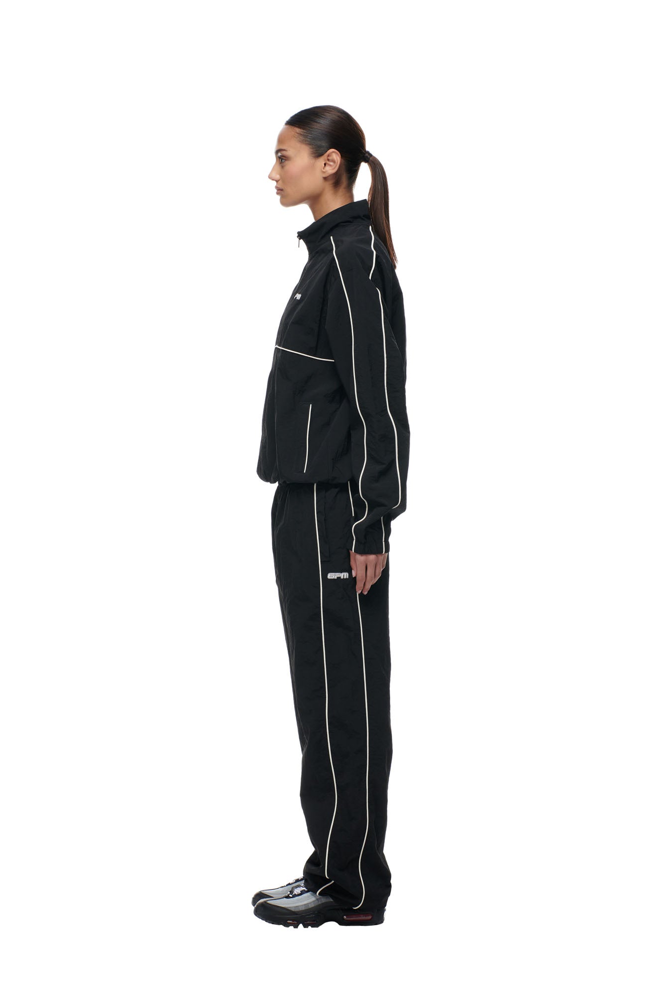 A person stands in profile against a plain white background, wearing the 6PM BF NYLON TRACKJACKET BLACK with white piping, black shoes, and their hair tied back in a ponytail.
