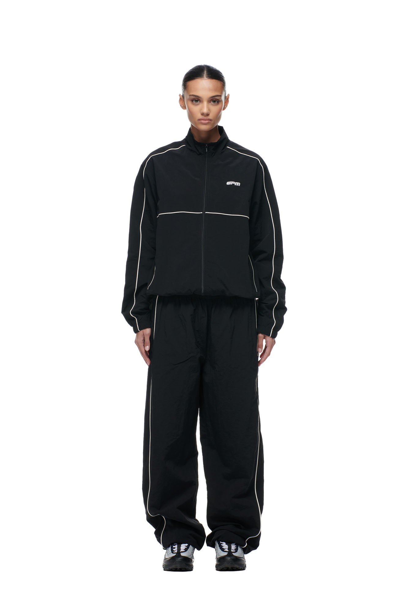 A person stands facing forward wearing the 6PM BF NYLON TRACKJACKET BLACK with matching black track pants and black sneakers. The zip-up jacket features white piping, and the background is plain white.
