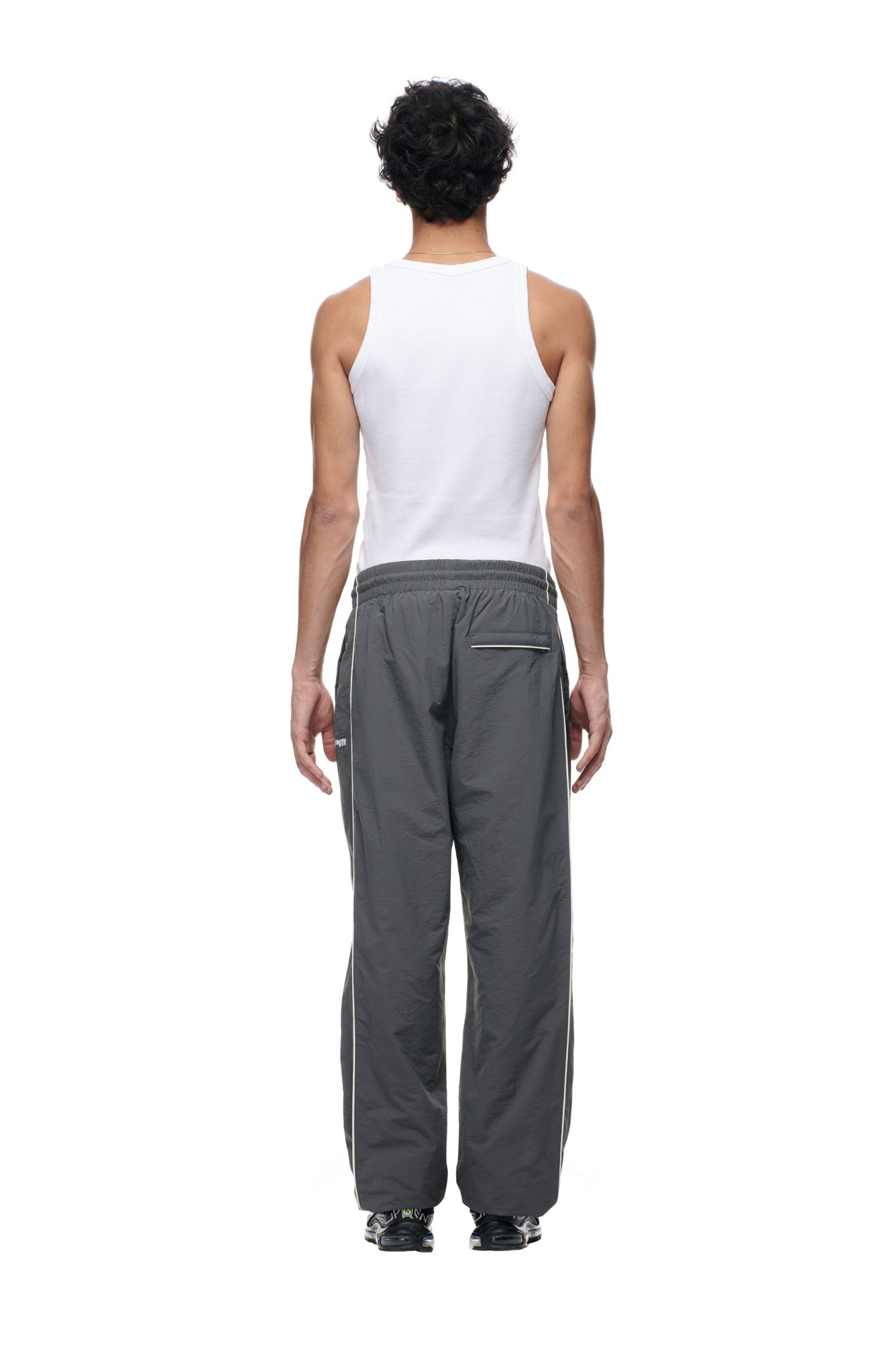 A person with short, dark, curly hair faces away from the camera, wearing a white sleeveless shirt, 6PM's BF NYLON TRACKPANTS ANTHRACITE, and black shoes against a plain white background.