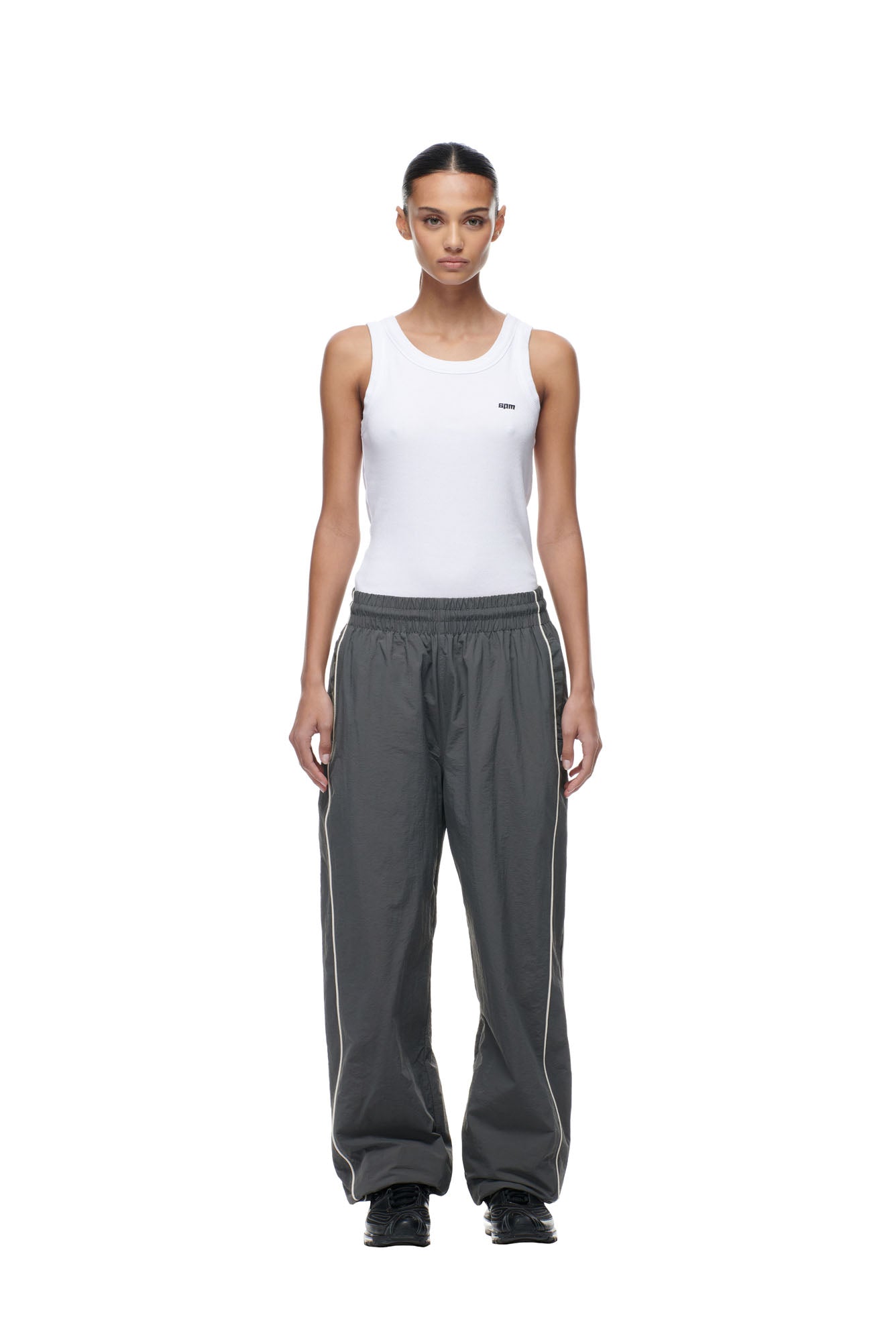 A woman stands facing forward against a white background, wearing 6PM’s BF NYLON TRACKPANTS ANTHRACITE paired with a white sleeveless tank top and black shoes.