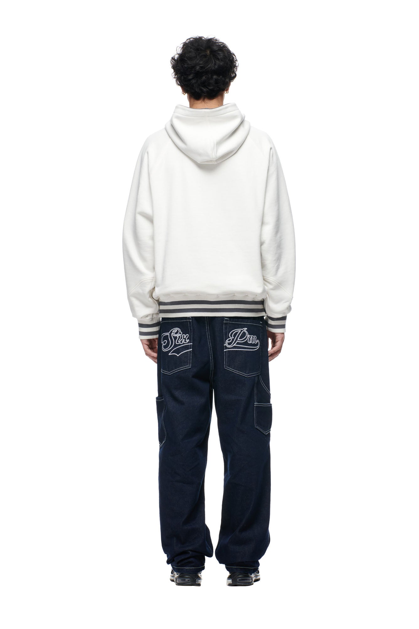 A person with short curly hair faces away, wearing the 6PM ONLY WEAR REVERSIBLE HOODIE in anthracite/cream and dark blue wide-leg jeans with white embroidery on the back pockets, against a plain white background.