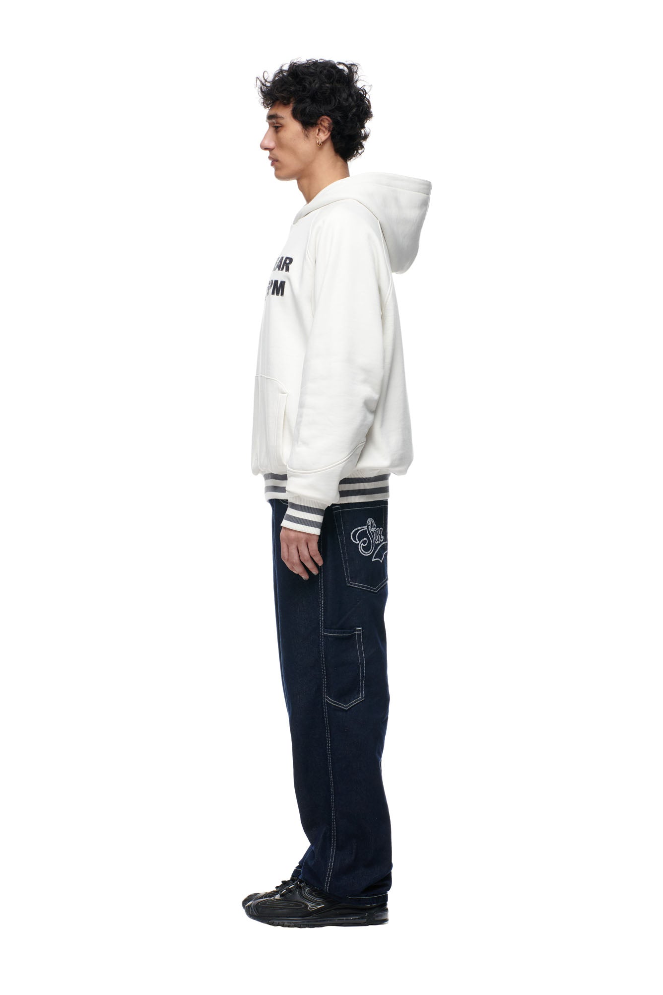 A young man stands in profile facing left, wearing the 6PM ONLY WEAR REVERSIBLE HOODIE in anthracite/cream, paired with dark blue jeans featuring a graphic back pocket and black sneakers, set against a plain white background.