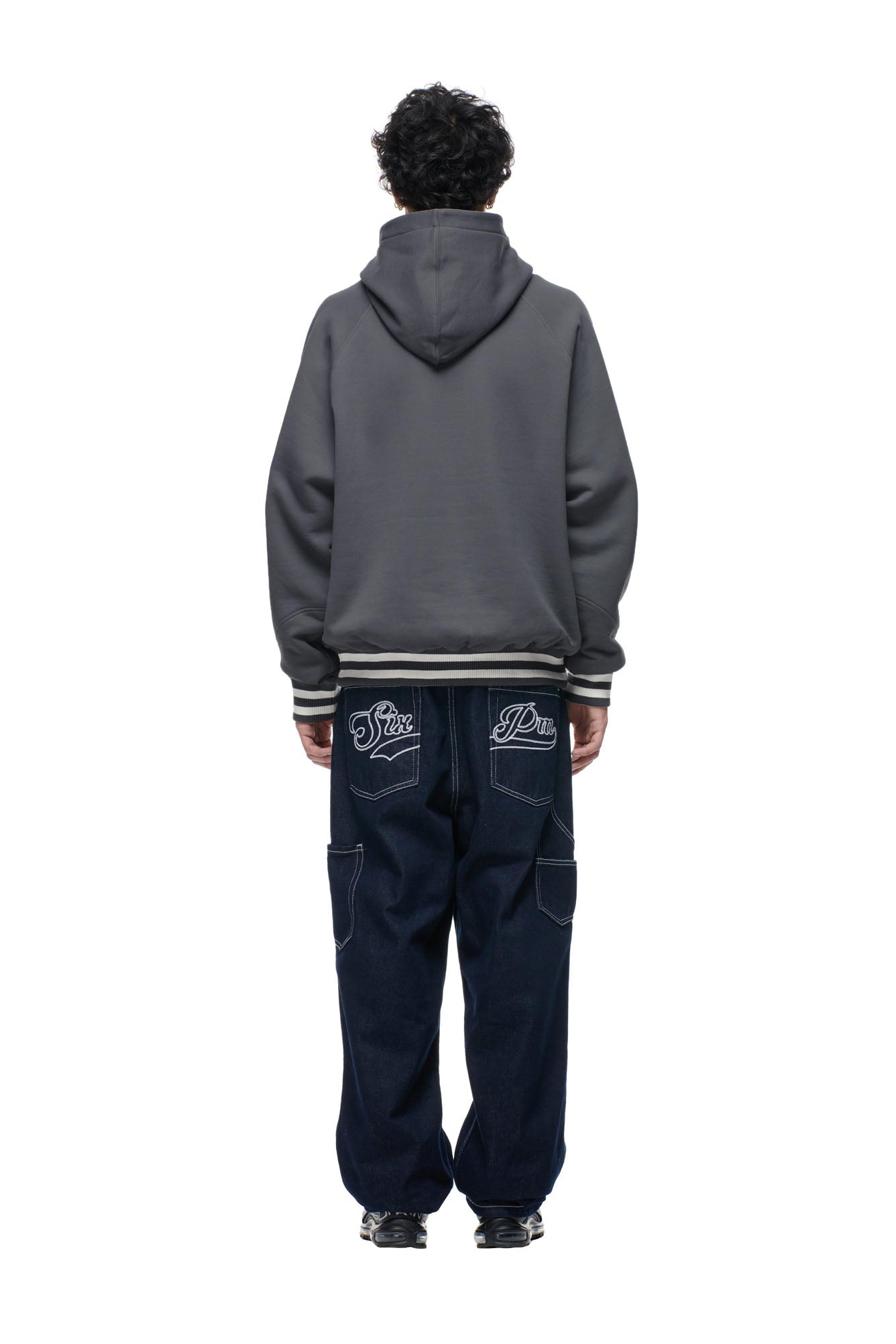 A person faces away from the camera wearing the 6PM ONLY WEAR REVERSIBLE HOODIE in anthracite/cream and loose dark blue jeans with white back pocket embroidery, set against a plain white background.