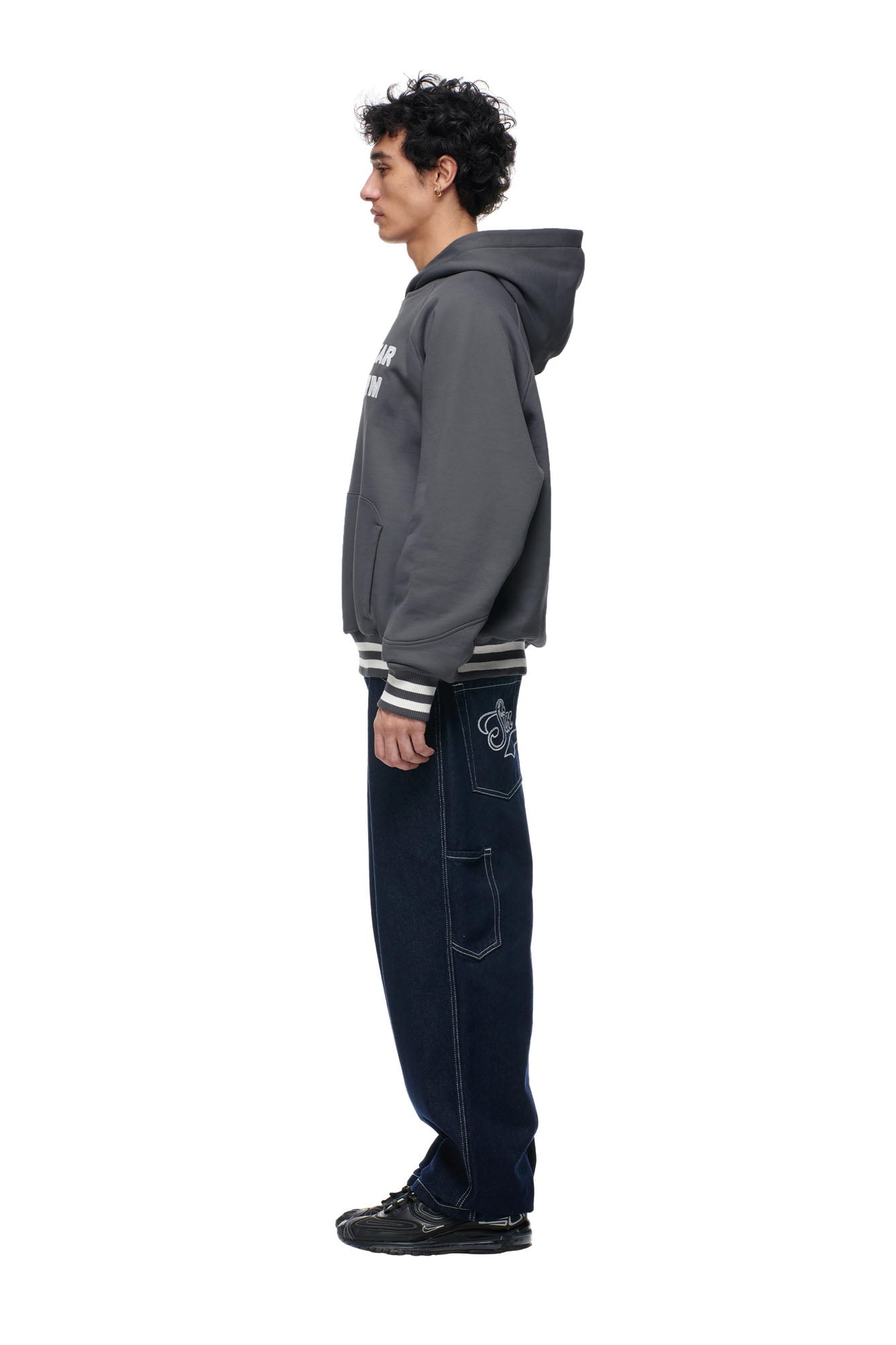 A man stands in profile against a white background, wearing the 6PM ONLY WEAR REVERSIBLE HOODIE in anthracite/cream, paired with loose dark blue jeans featuring side designs and black sneakers.