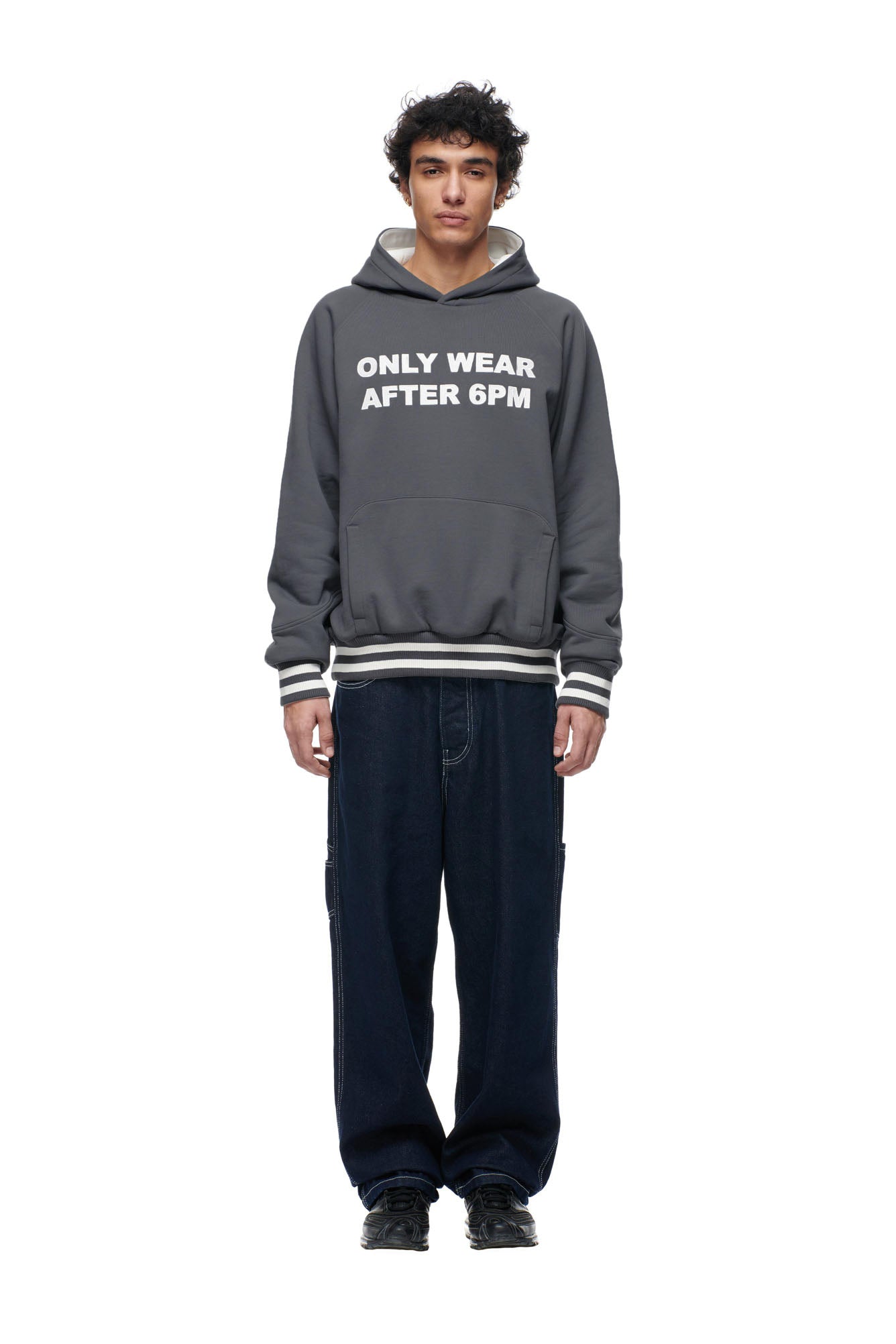 A person with curly hair wears the 6PM ONLY WEAR REVERSIBLE HOODIE in anthracite/cream and loose dark pants, standing facing forward against a plain white background.