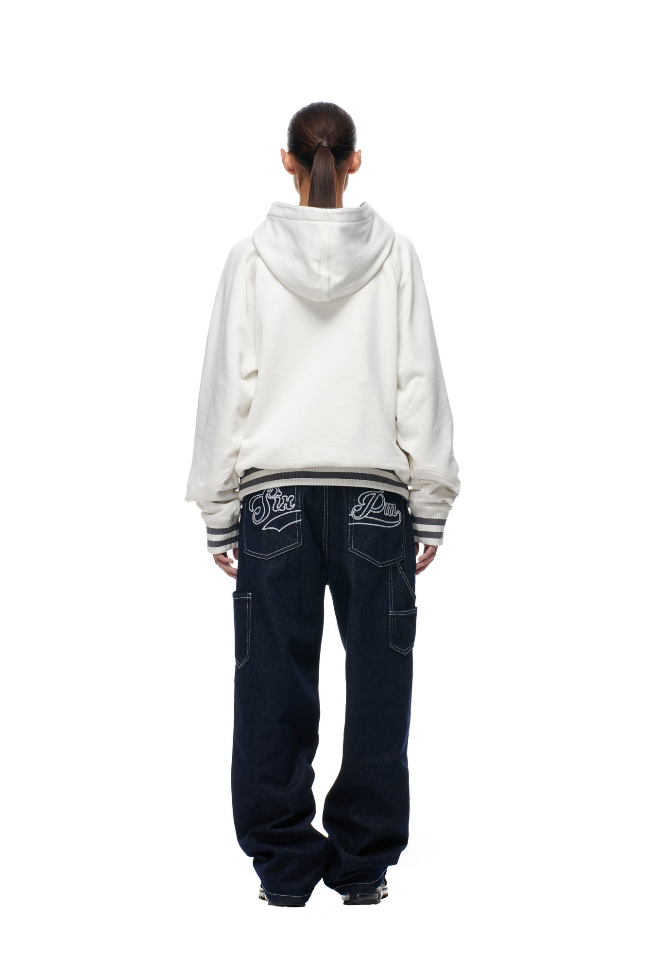 A person stands facing away wearing the 6PM ONLY WEAR REVERSIBLE HOODIE in anthracite/cream and baggy dark blue jeans with decorative stitching and writing on the back pockets, set against a plain white background.