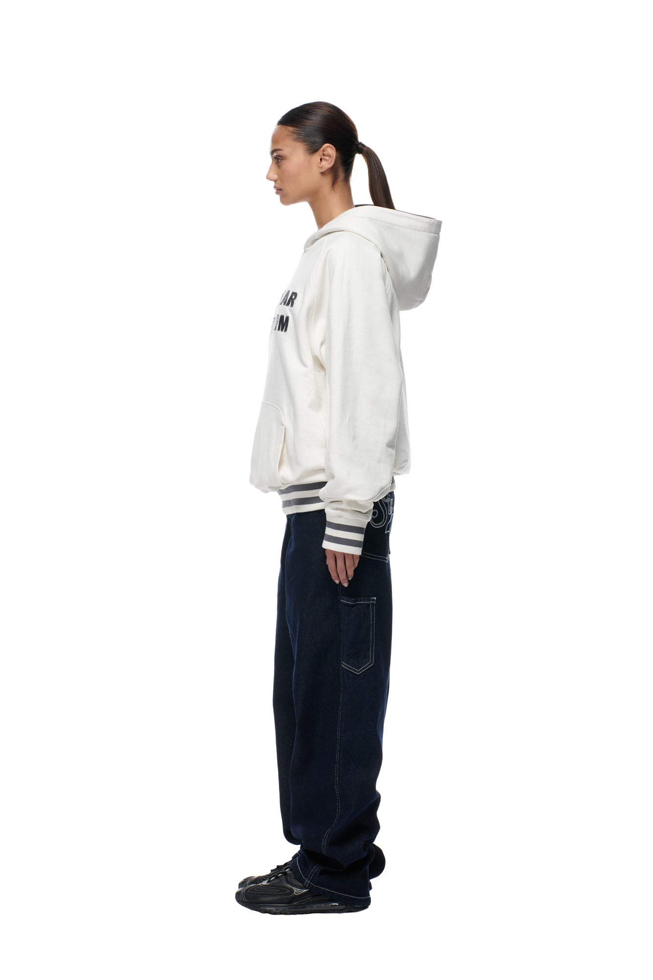 A person stands in profile against a white background, wearing the 6PM ONLY WEAR REVERSIBLE HOODIE ANTHRACITE/CREAM, dark loose jeans, and black sneakers. Their hair is tied back in a low ponytail.