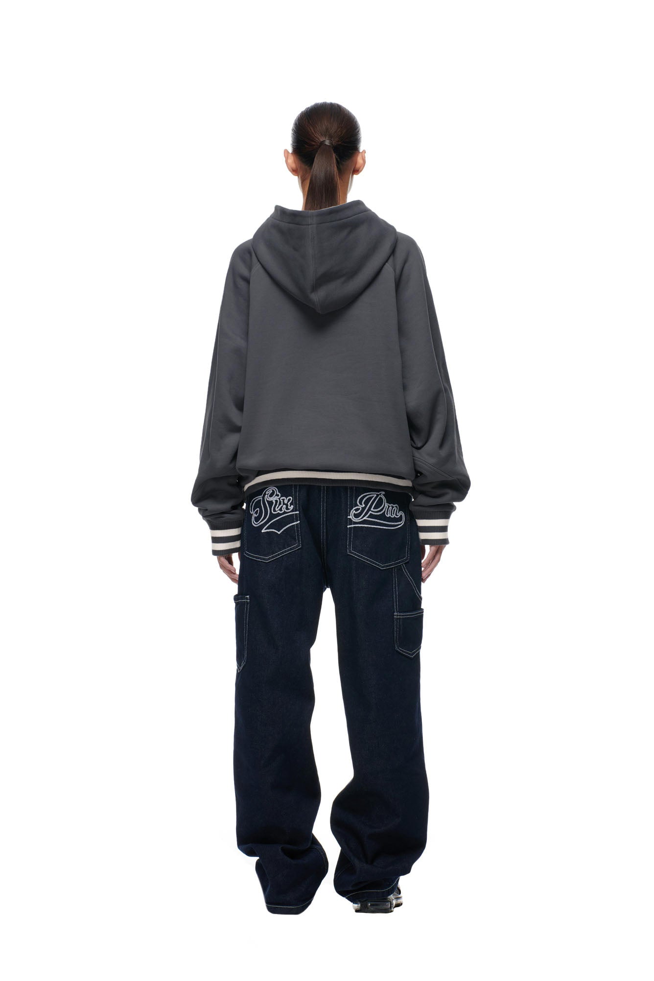 A person with long hair in a ponytail is seen from behind, wearing the 6PM ONLY WEAR REVERSIBLE HOODIE in anthracite/cream and loose dark jeans featuring white stitching and embroidered back pocket designs.